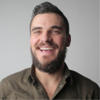 Smiling man with dark hair and beard wearing an olive green shirt against a light gray background.