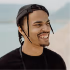 Smiling young man with braided hair wearing a backward black cap and black shirt outdoors near water.
