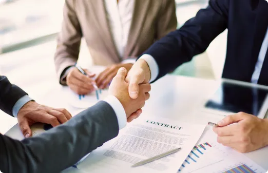 Two businesspeople shaking hands over a signed contract at a desk with documents and a tablet.