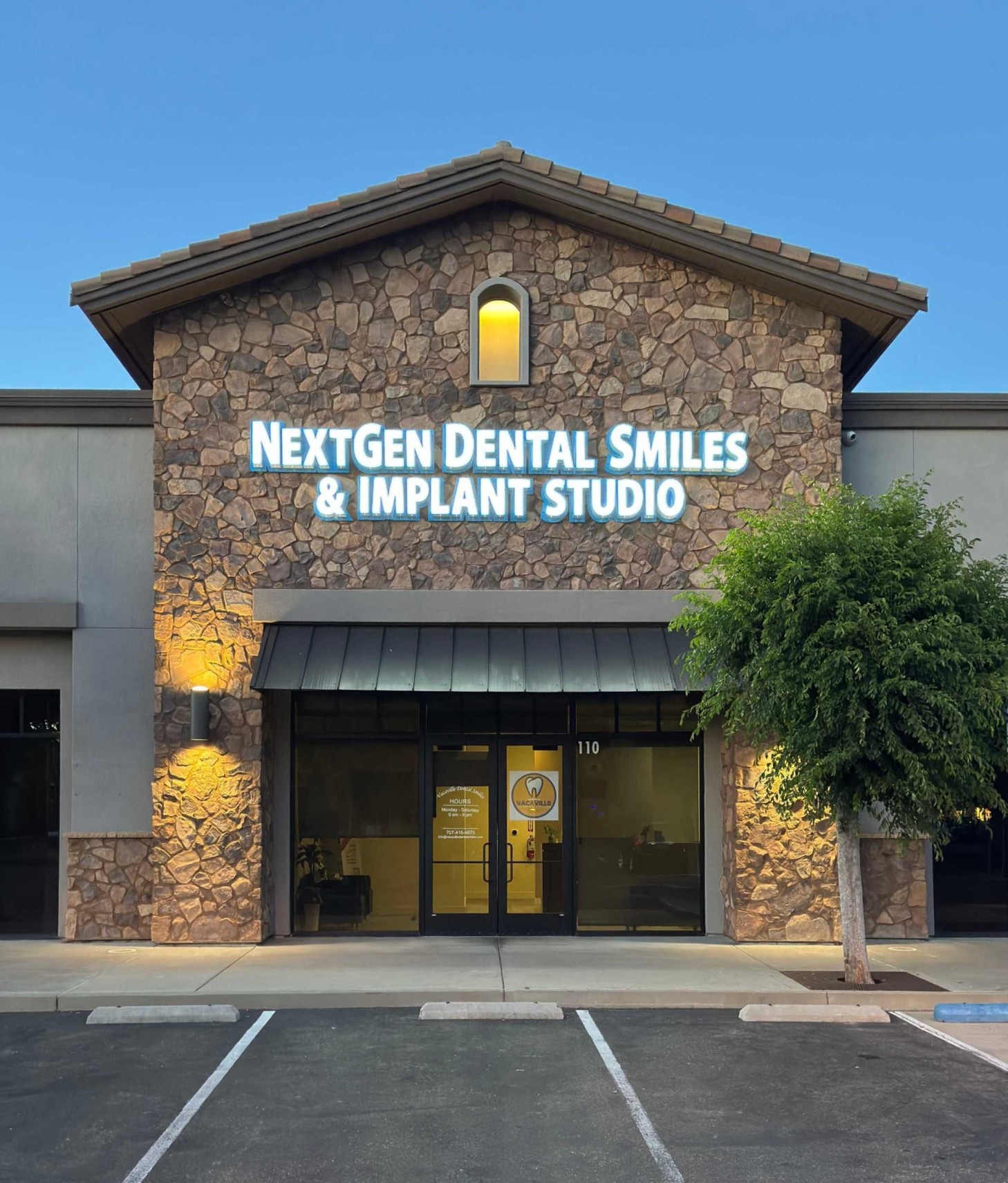 Stone facade of NextGen Dental Smiles & Implant Studio storefront with glass doors and a small tree to the right.