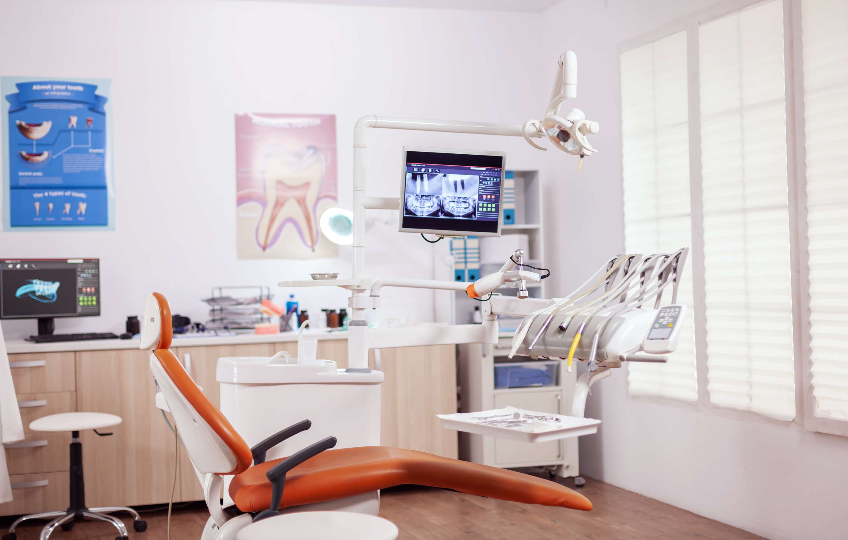 Modern dental clinic with orange dental chair, digital X-ray screen, dental tools, and educational posters on the wall.
