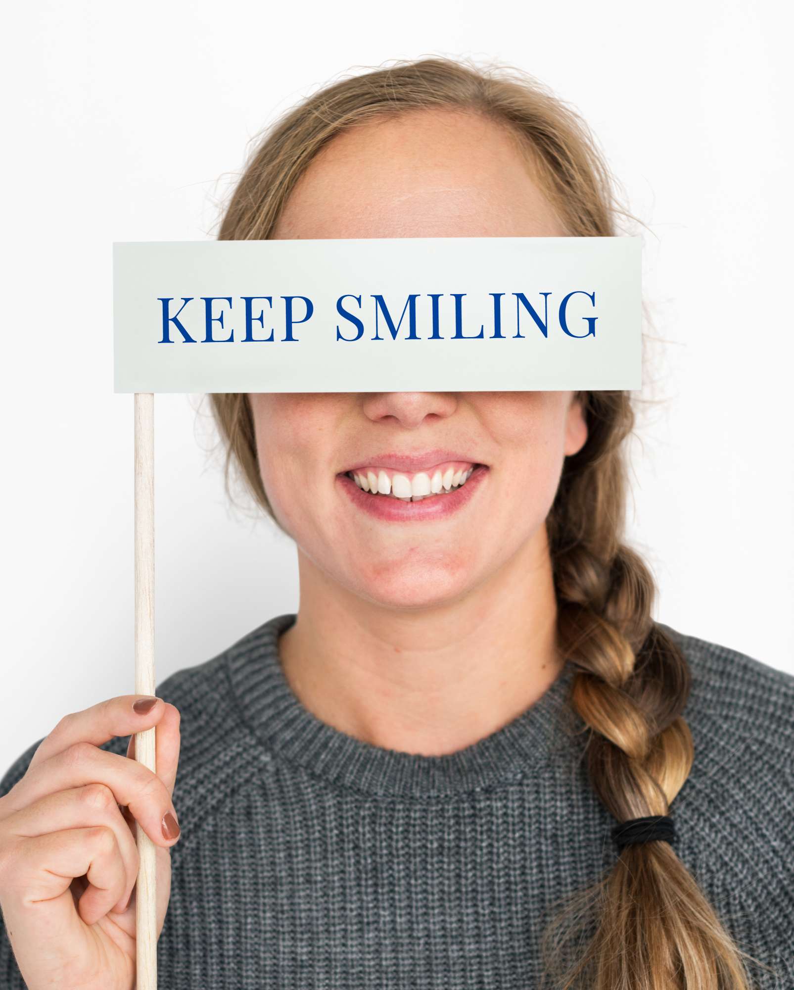 Woman with braided hair holding a sign that reads 'KEEP SMILING' covering her eyes and smiling.
