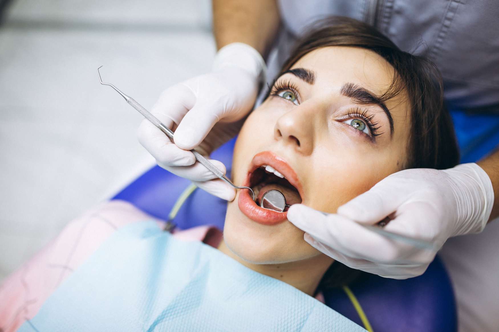 Woman with green eyes receiving a dental checkup with mouth open while dentist wearing gloves uses dental tools.