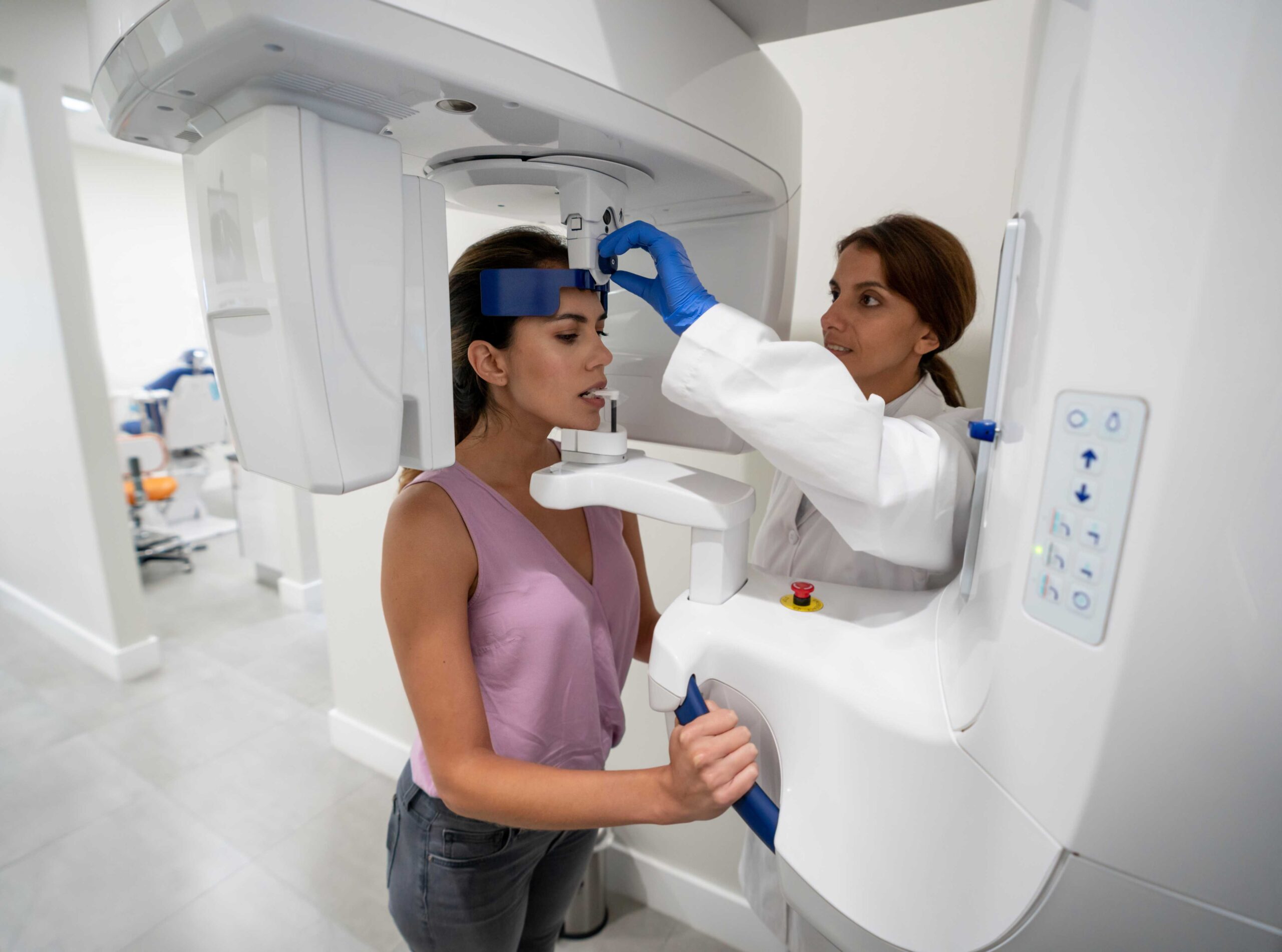 Female patient standing in a dental X-ray machine while a technician adjusts the device.