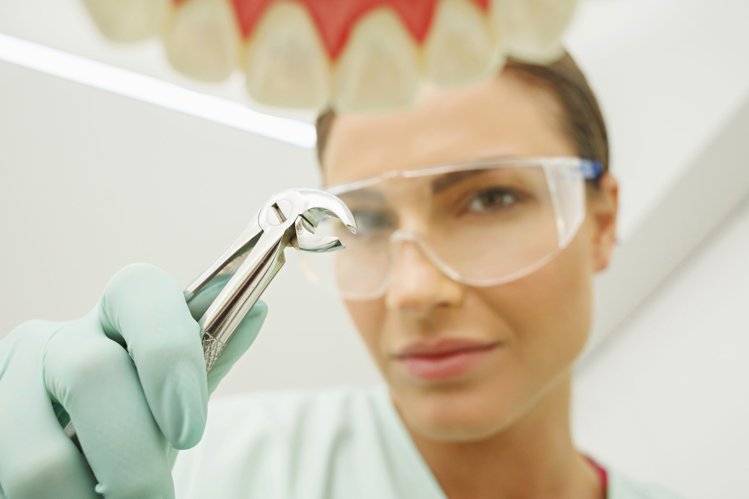 Dentist wearing safety glasses and gloves holding dental extraction forceps near an open mouth.