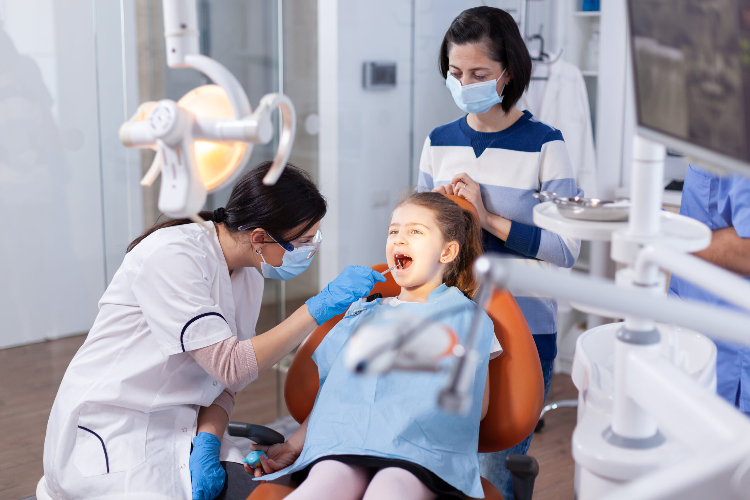Dentist wearing mask and gloves examining a young girl’s mouth while her mother watches in a dental clinic.