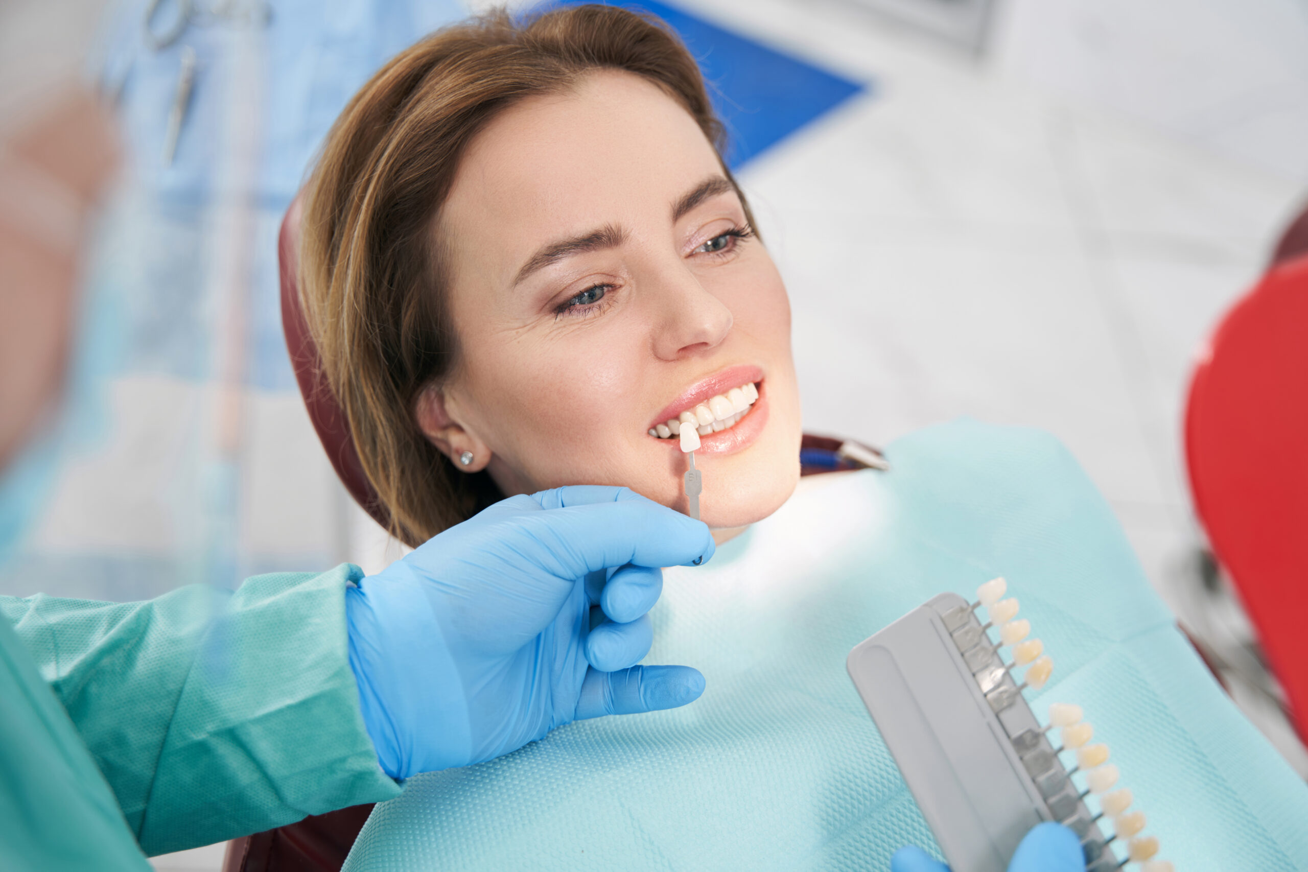 Dentist wearing blue gloves matching a tooth shade guide to a smiling female patient's teeth in a dental chair.