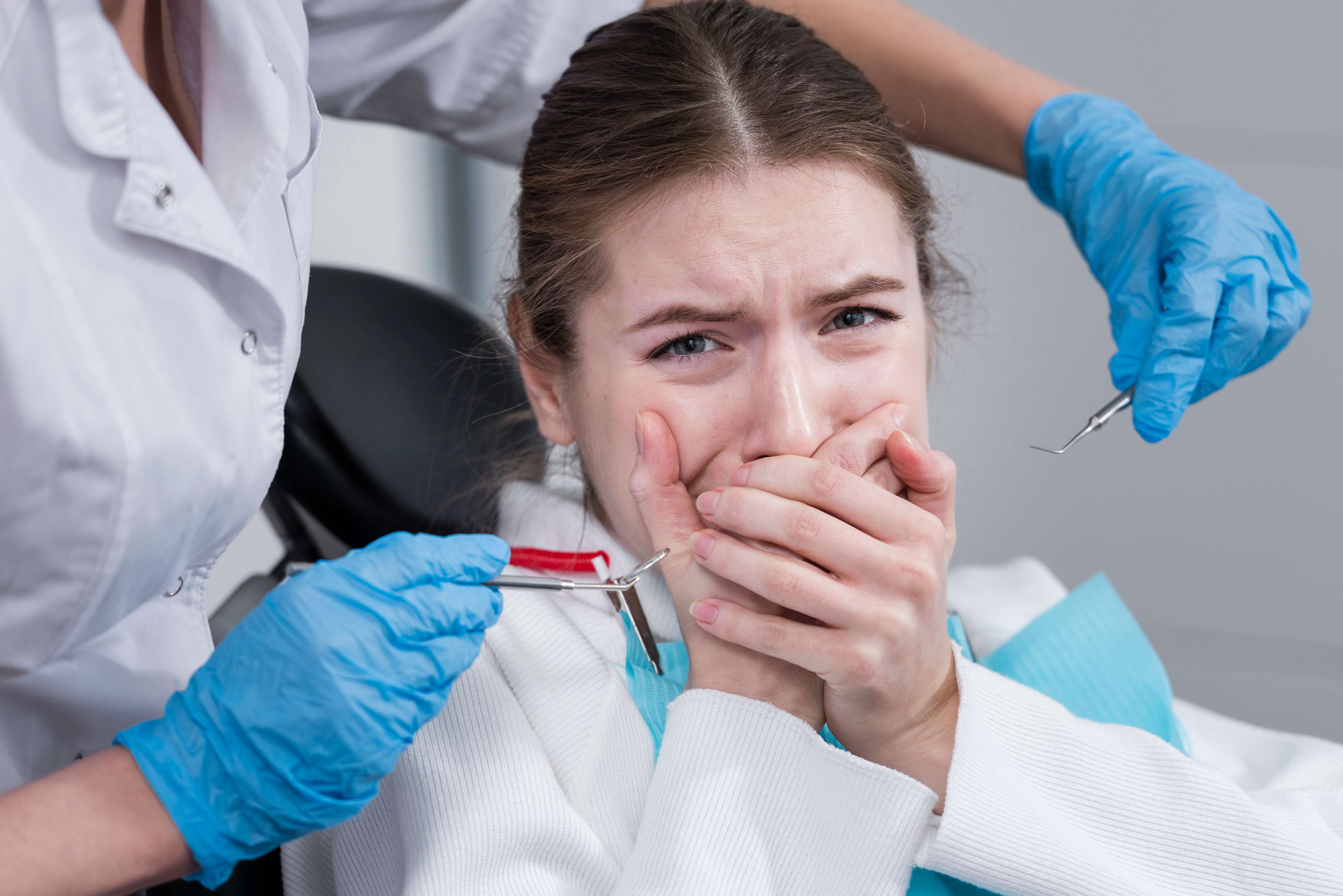 Anxious woman covering her mouth in a dental chair as a dentist wearing blue gloves holds dental tools near her.