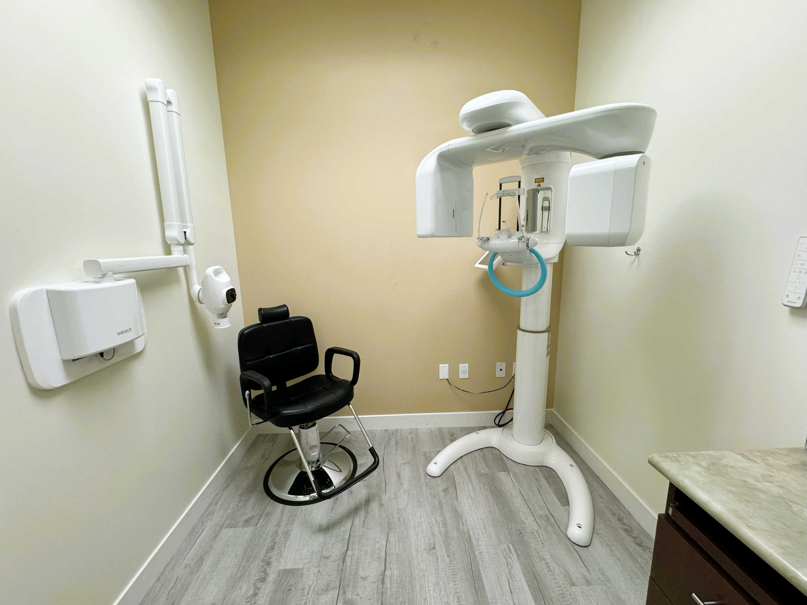 Dental x-ray room with a black patient chair, white x-ray machine, and light-colored walls and flooring.