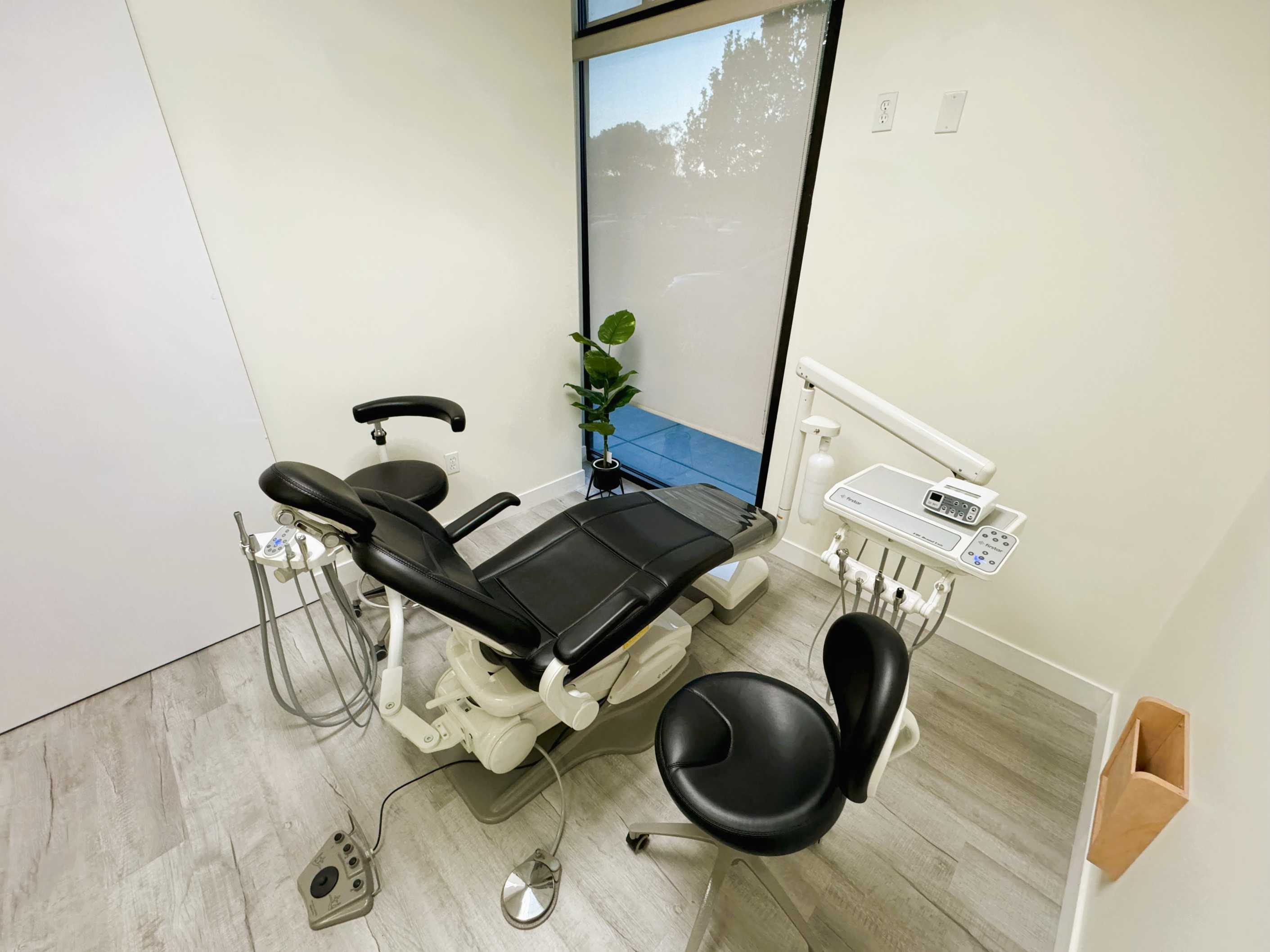 Empty modern dental clinic room with a black dental chair, dentist stool, and dental instruments near a window with a roller shade partially down.