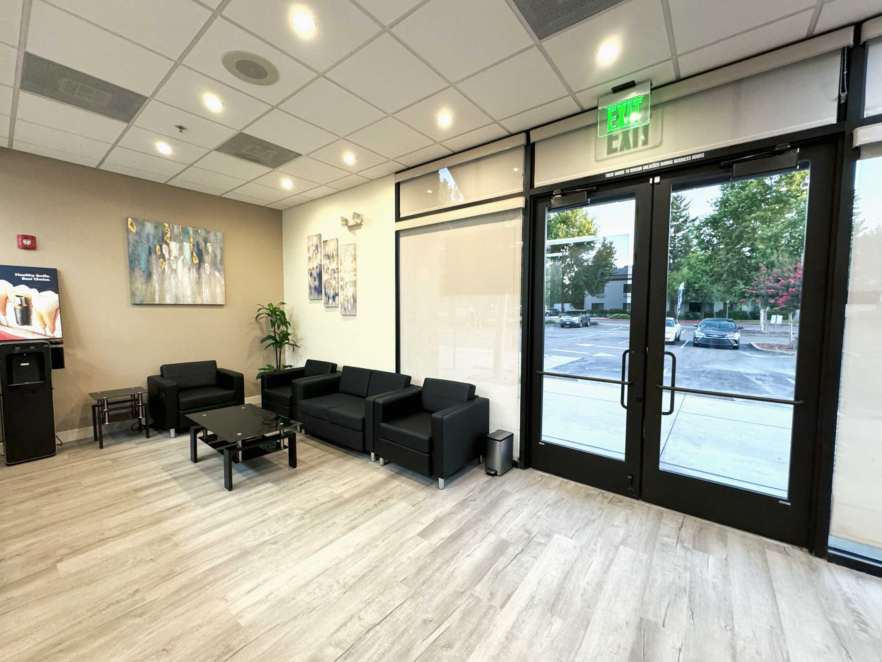 Modern waiting room with black leather sofas, glass coffee table, wall art, and glass exit doors showing a parking lot outside.