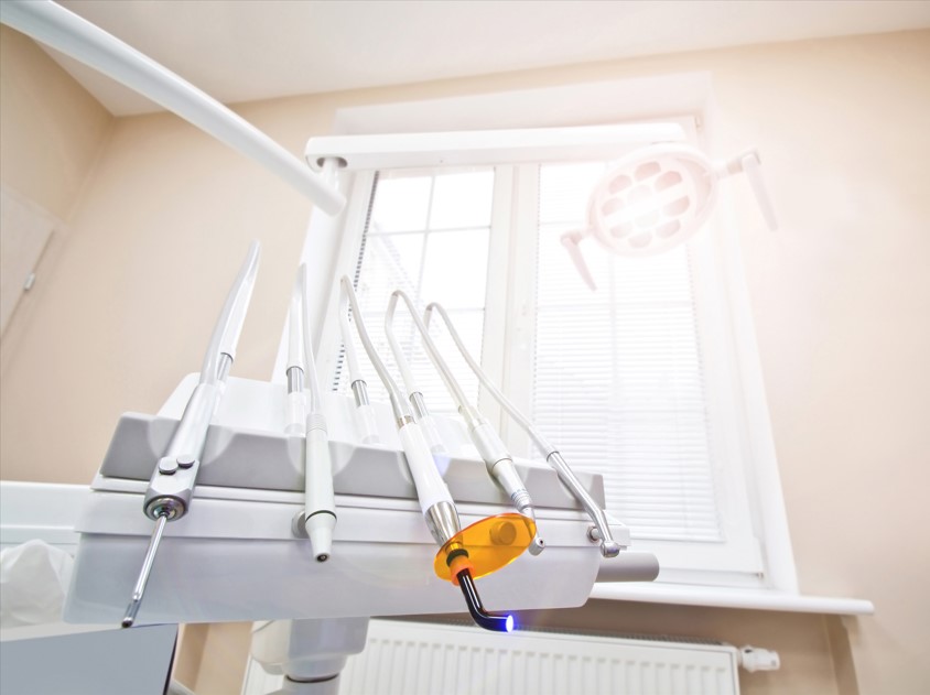 Close-up of dental instruments on a white holder with a lit blue dental curing light in a bright clinic room.