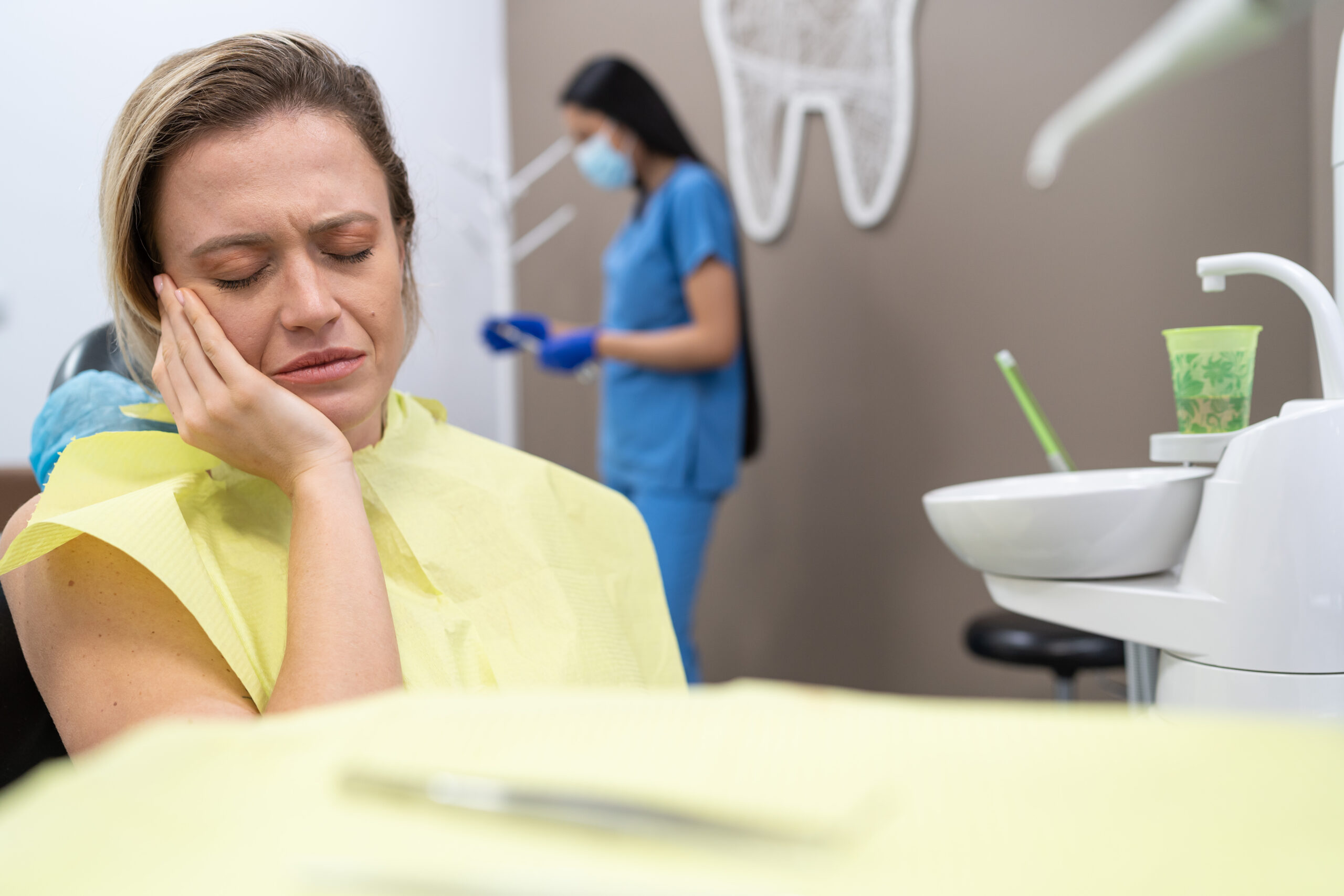 Woman in dental chair wearing yellow bib, holding her cheek in pain with a dental assistant in blue scrubs and mask in the background.