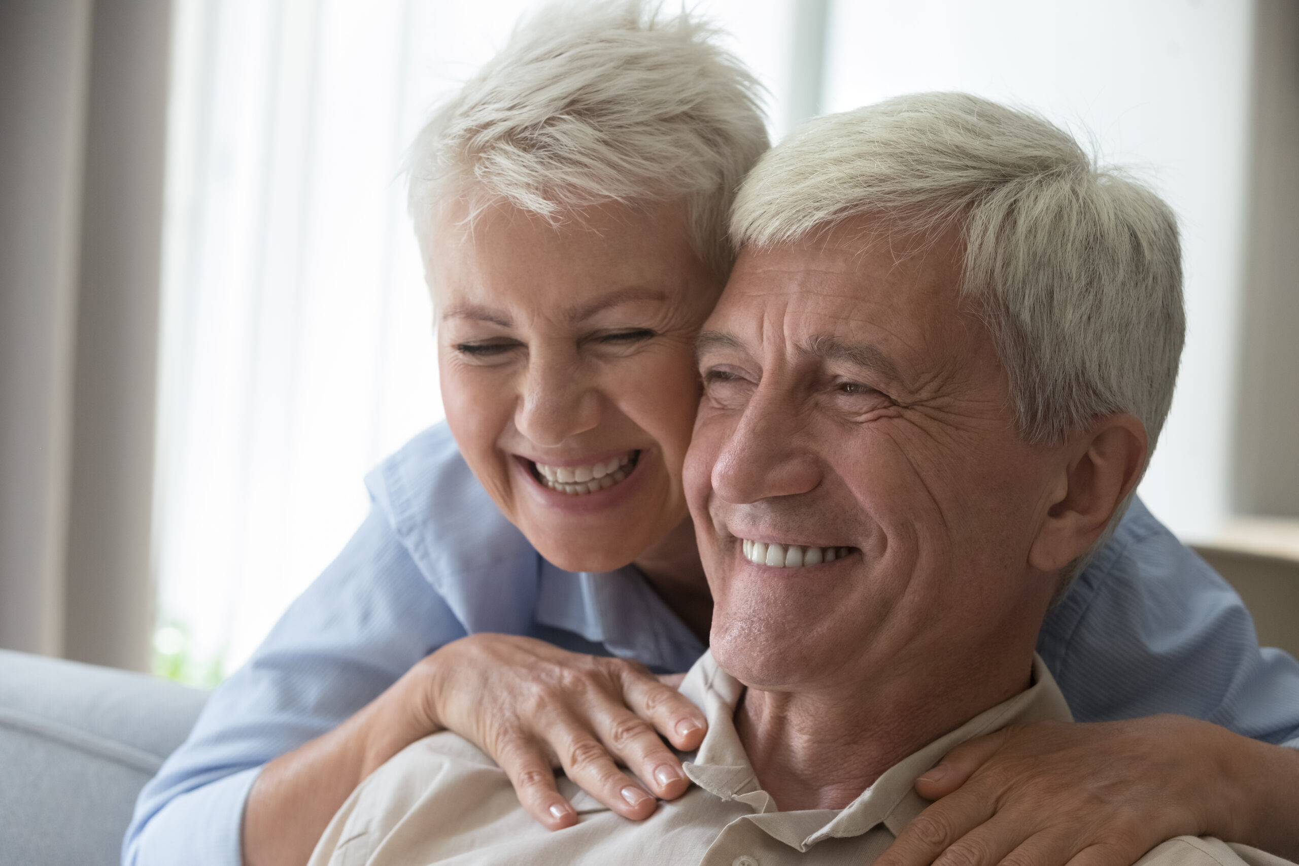 Smiling elderly woman with short white hair hugging a smiling elderly man with gray hair from behind.