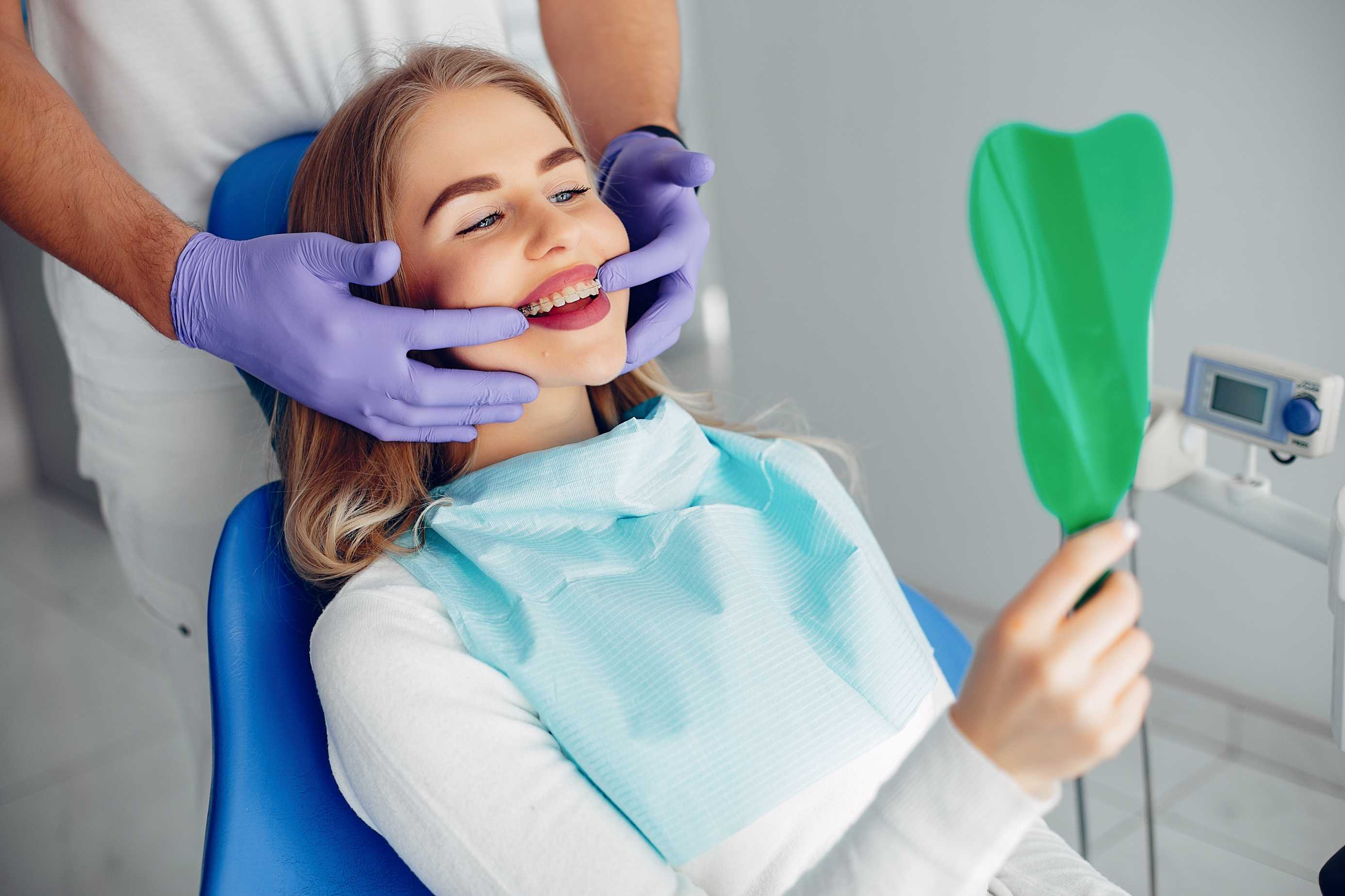 Woman with braces smiling in a dental chair holding a green tooth-shaped mirror, while a dentist in purple gloves examines her teeth.