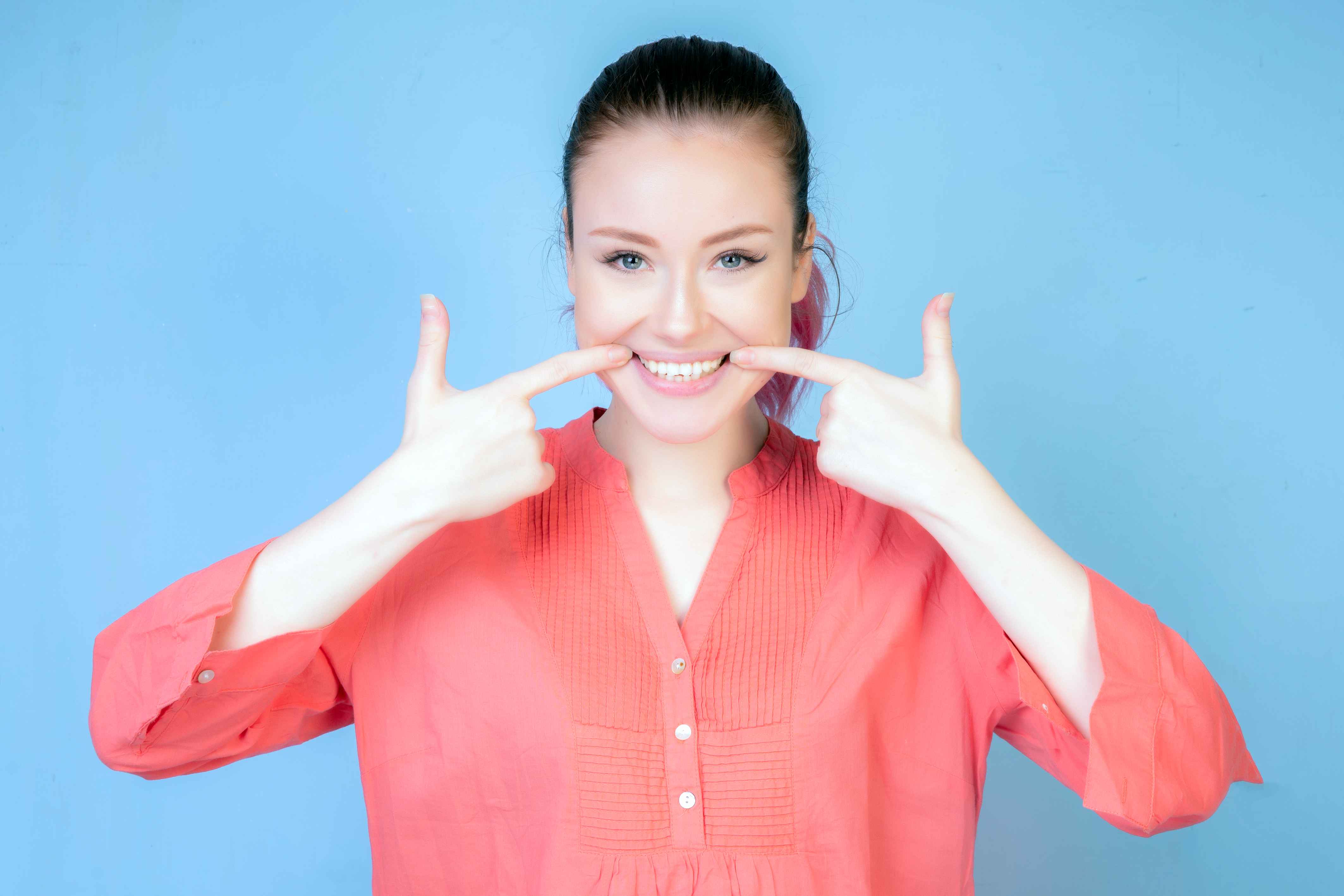 Smiling woman in coral shirt pointing to her white teeth with both index fingers against a blue background.