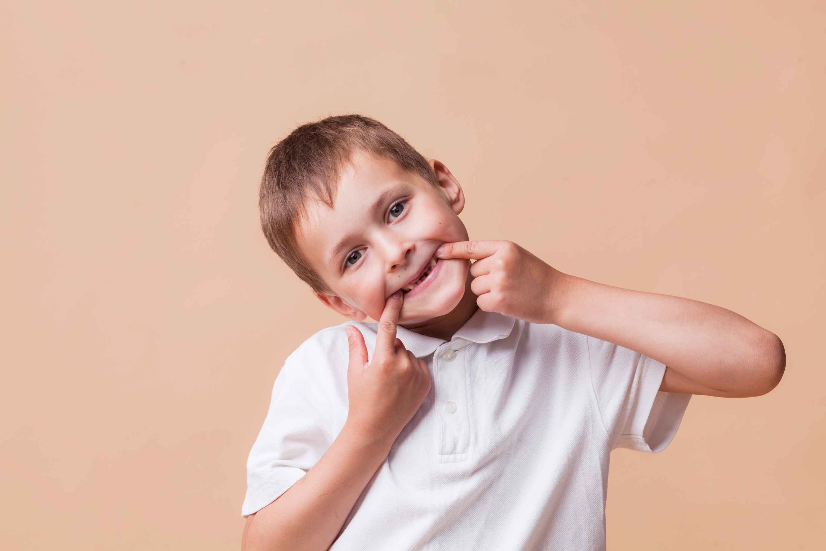 Young boy in white shirt smiling and pointing at his missing front teeth against a beige background.