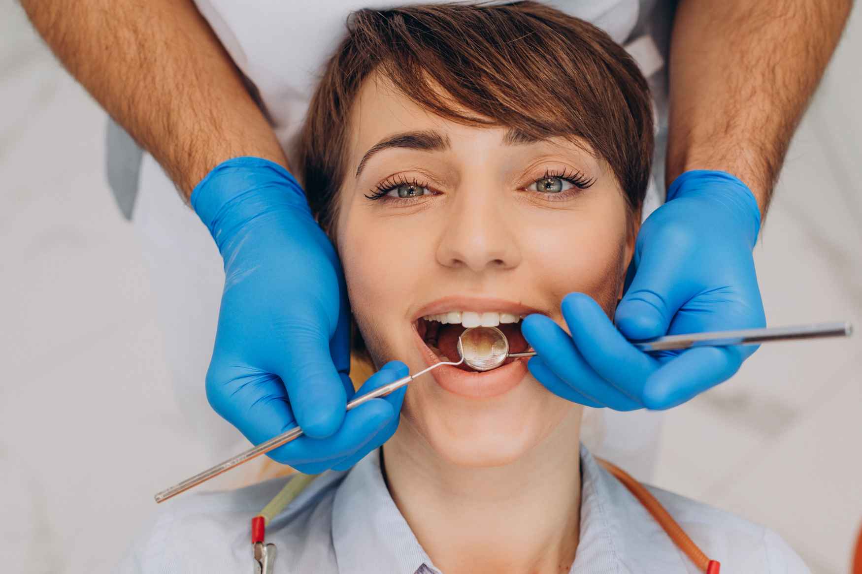 Dentist wearing blue gloves examining a woman's teeth using dental mirror and probe.