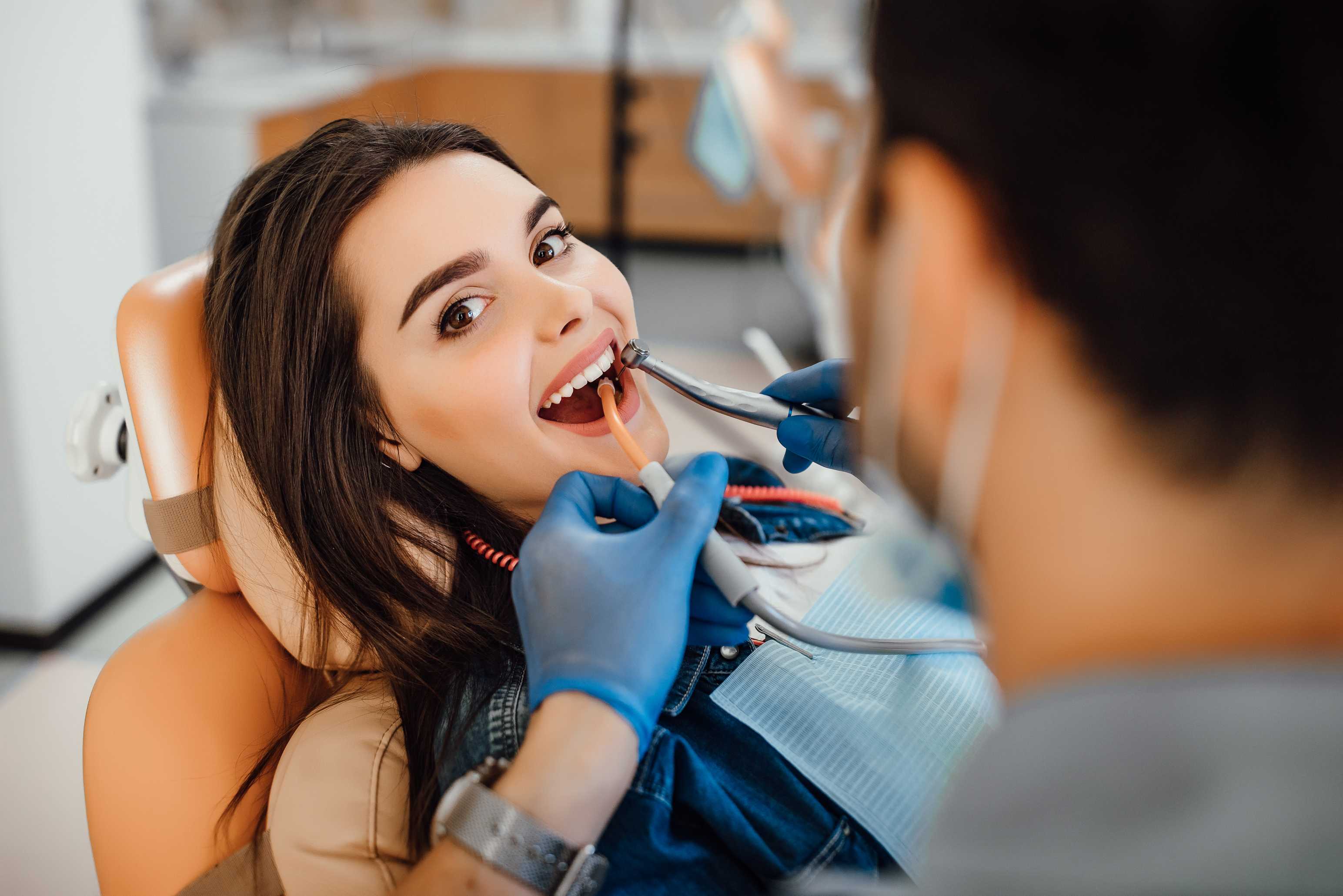 Female patient with dark hair smiling in dental chair while dentist wearing blue gloves works on her teeth with dental tools.