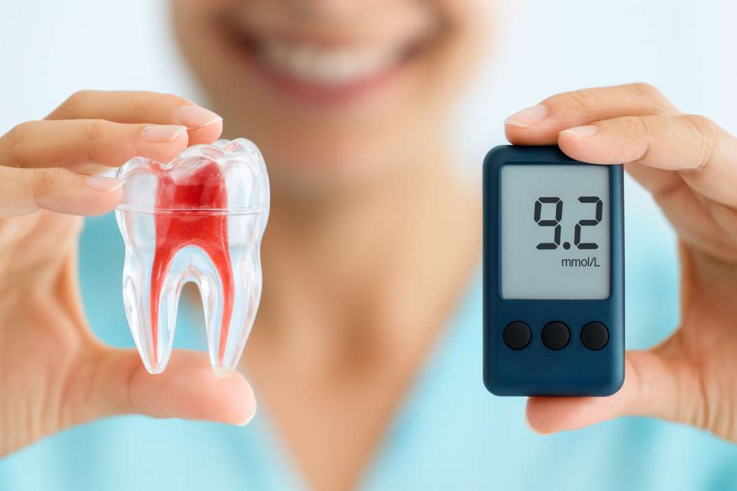 Person holding a clear model of a tooth with red root detail in one hand and a blood glucose meter showing 9.2 mmol/L in the other.