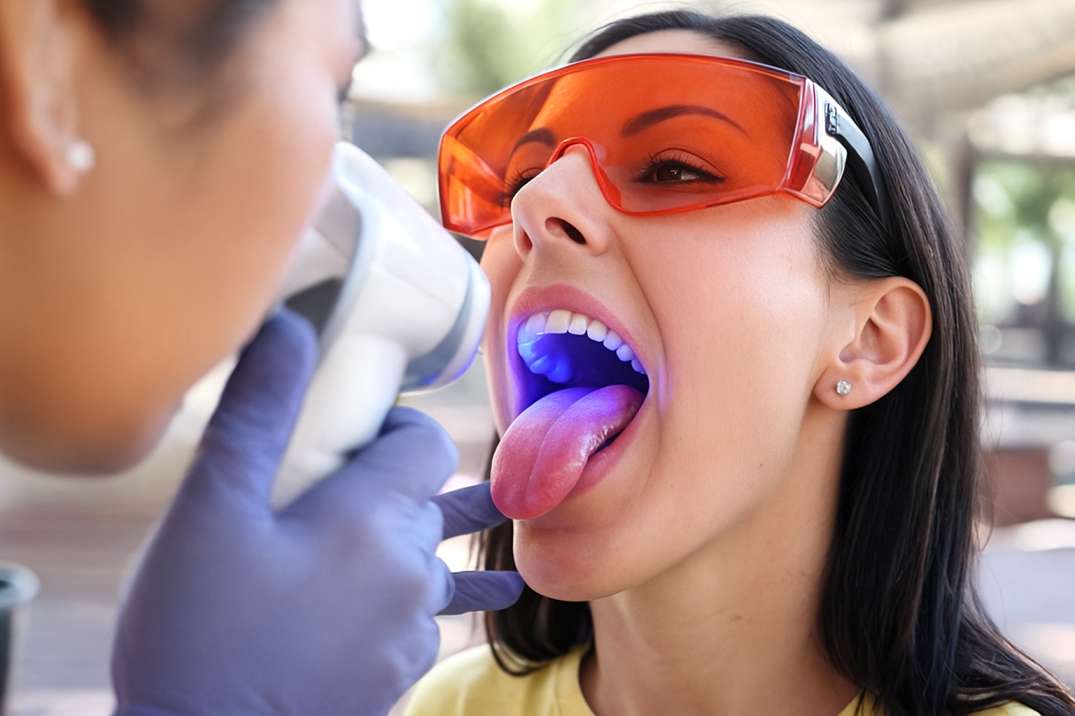 A woman wearing orange protective glasses has her mouth open with tongue out while a dental professional uses a handheld blue light device to examine her oral cavity.