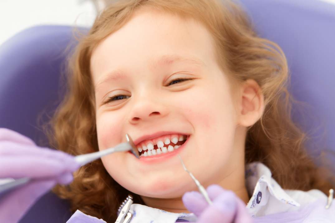 Smiling young girl with curly hair sitting in a dental chair during a teeth cleaning.