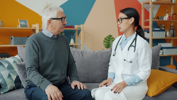 Young woman medic is talking to old man touching his shoulder expressing support visiting patient in apartment. Communication and medicare concept.