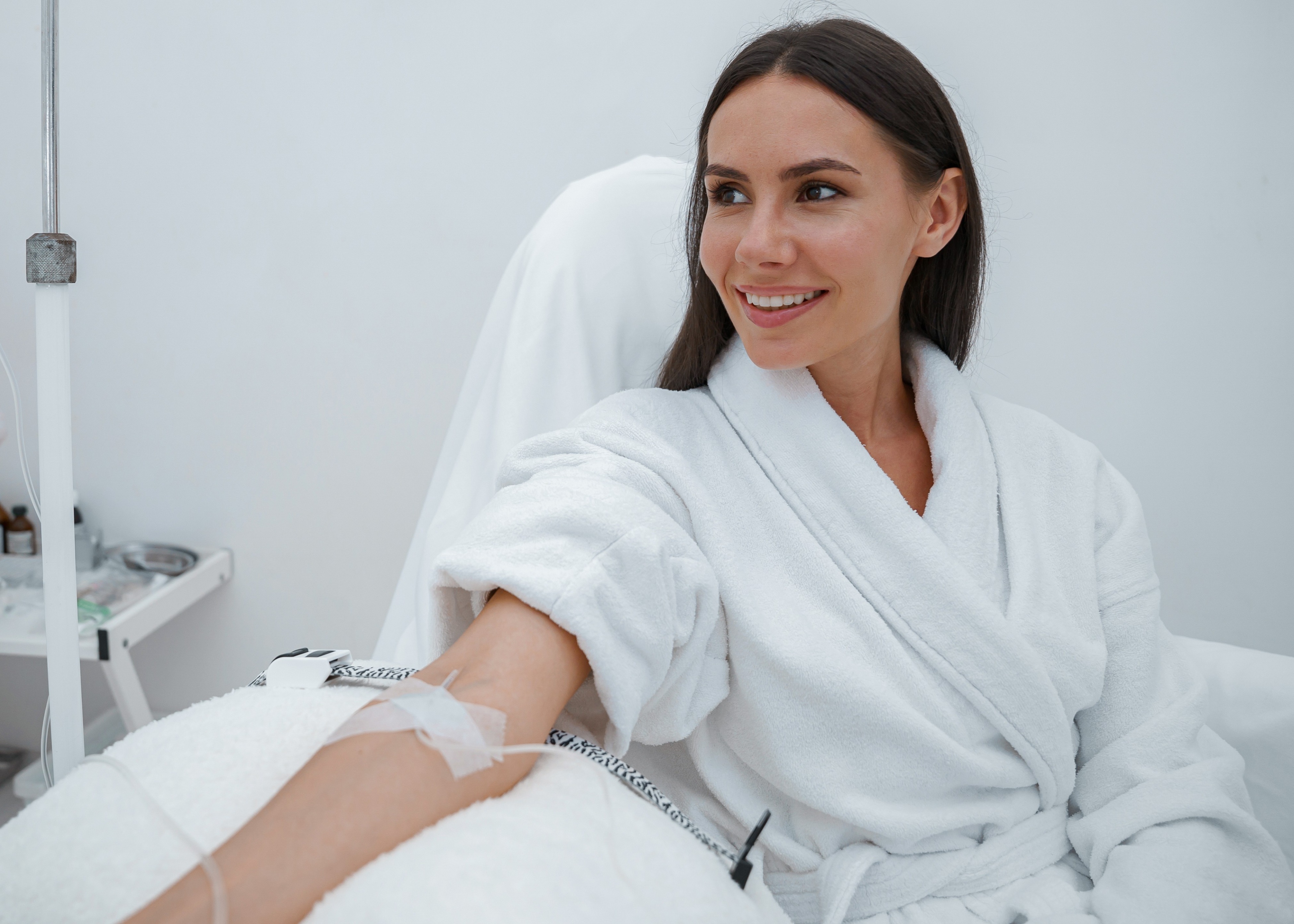 Smiling woman in a white robe receiving intravenous therapy while sitting in a medical chair.