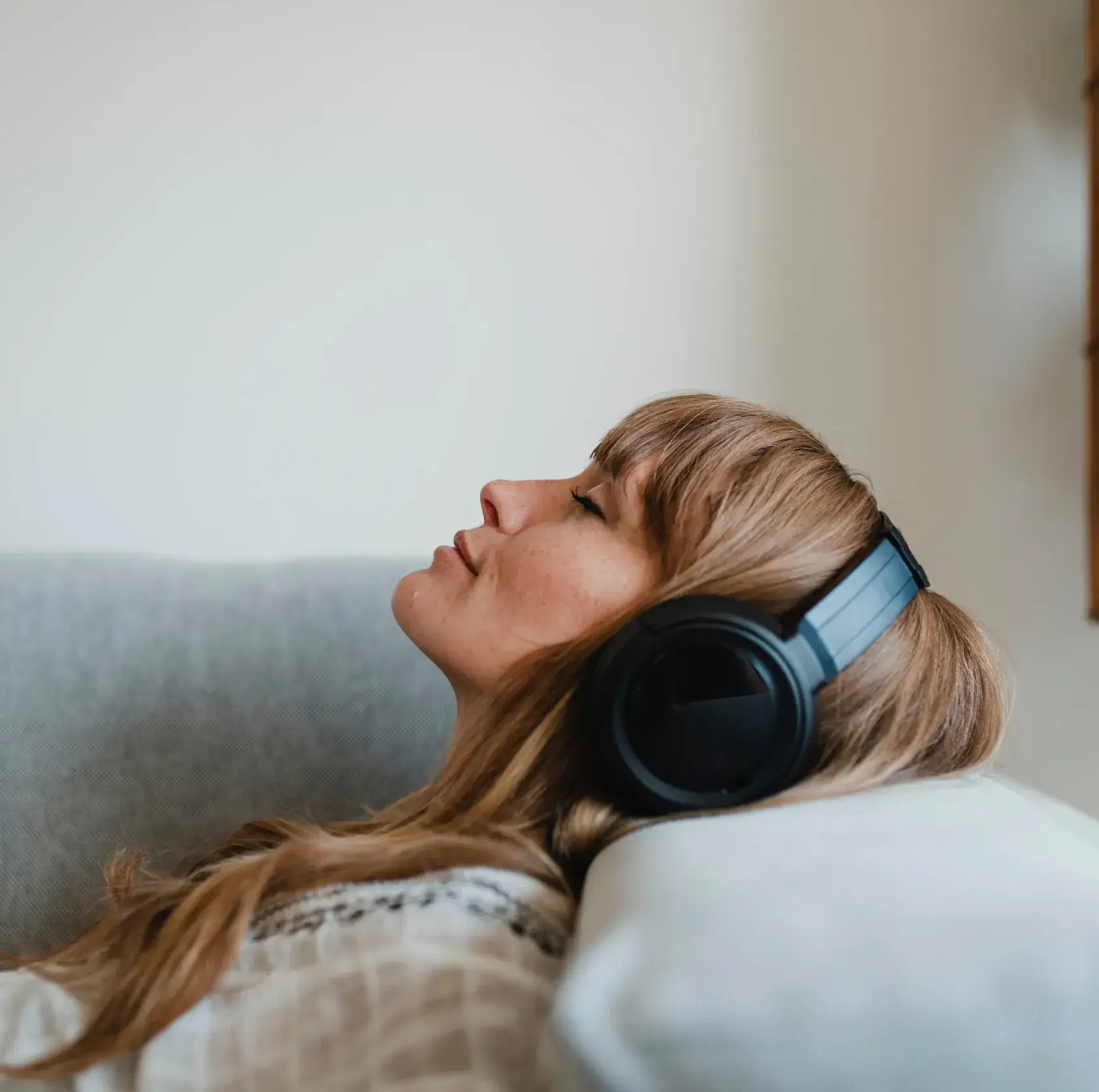 Woman with closed eyes wearing black headphones, reclining on a gray couch.