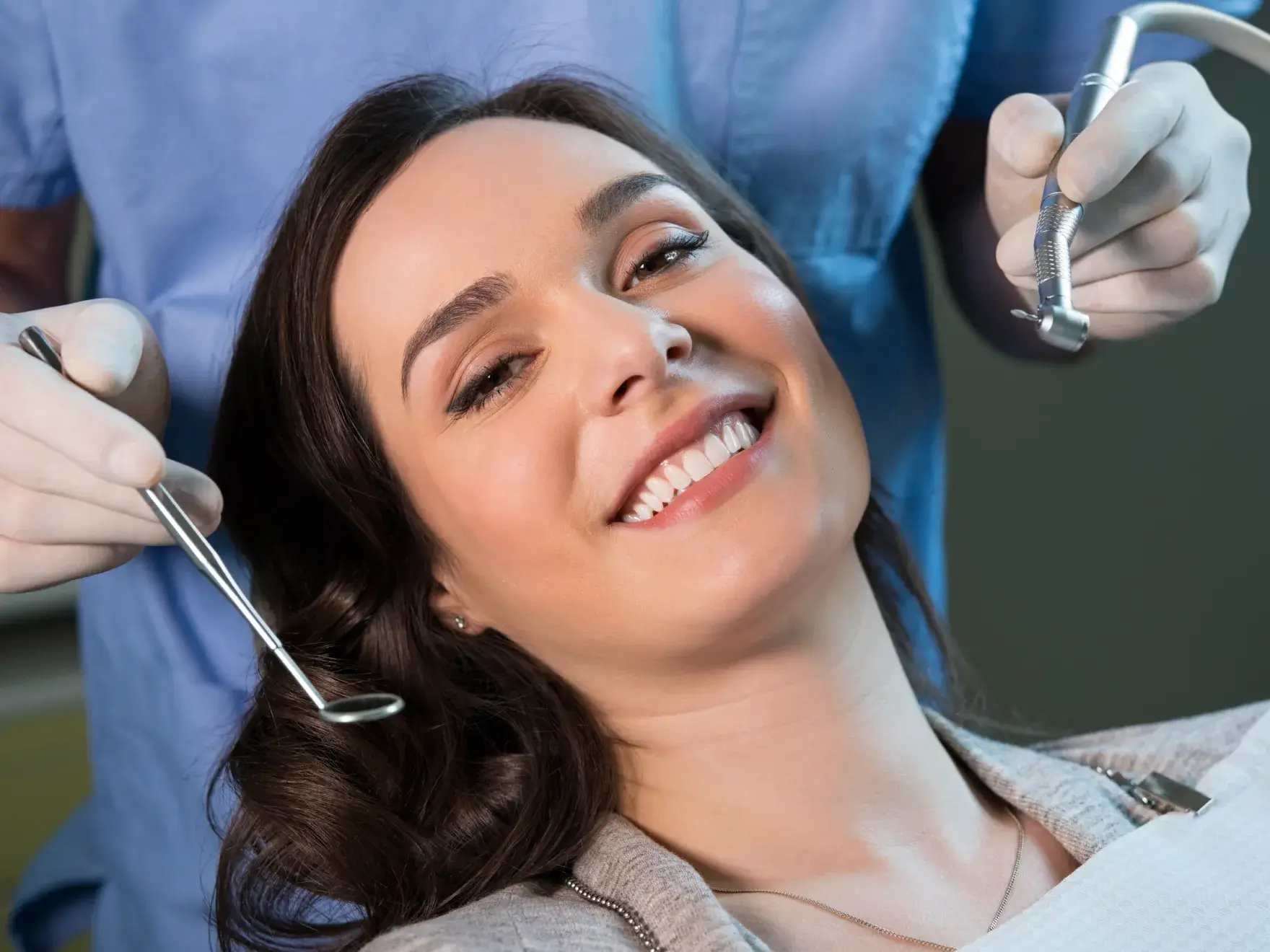 Smiling woman reclining in dental chair with dentist holding dental mirror and drill.