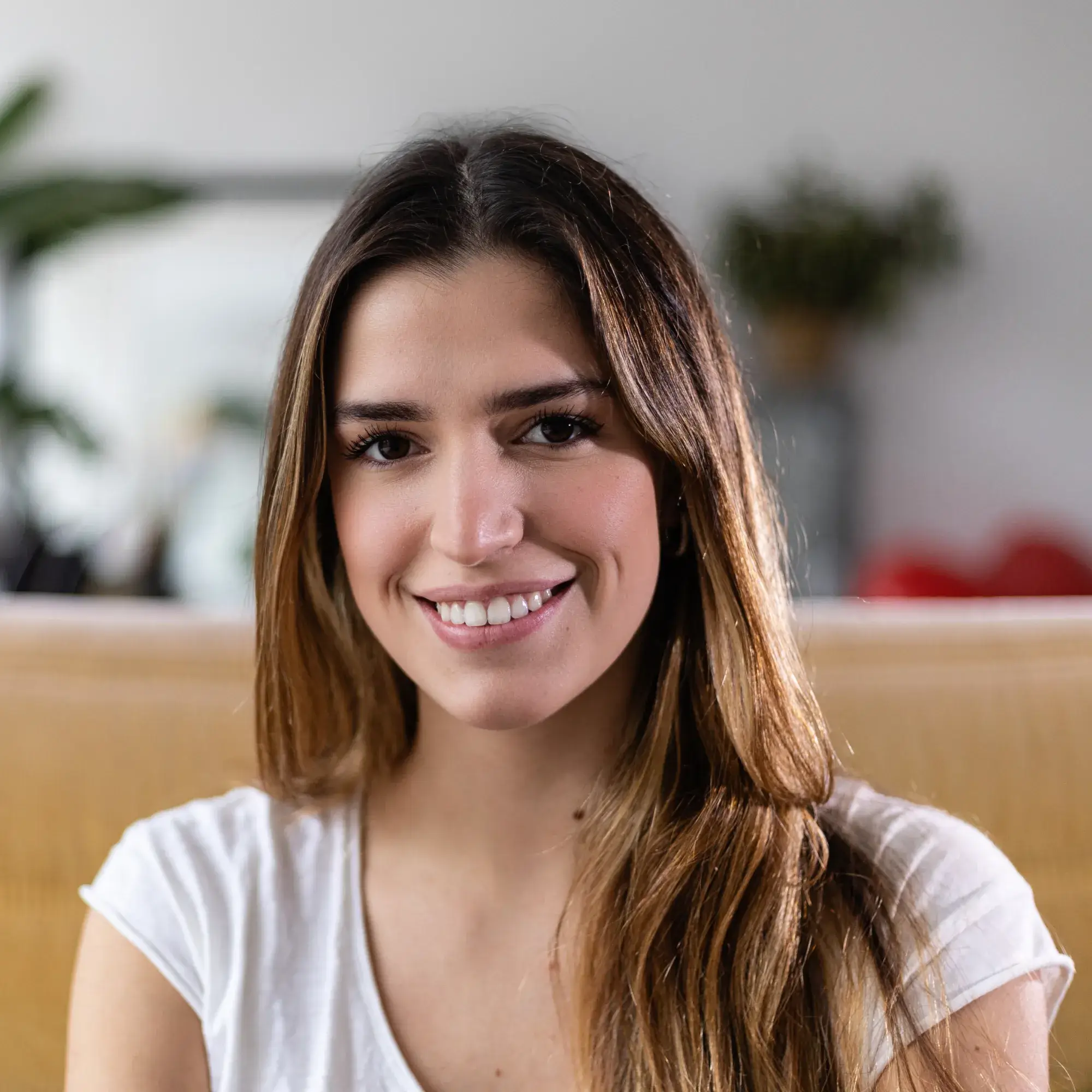 Smiling woman with long brown hair wearing a white shirt sitting indoors on a sofa.