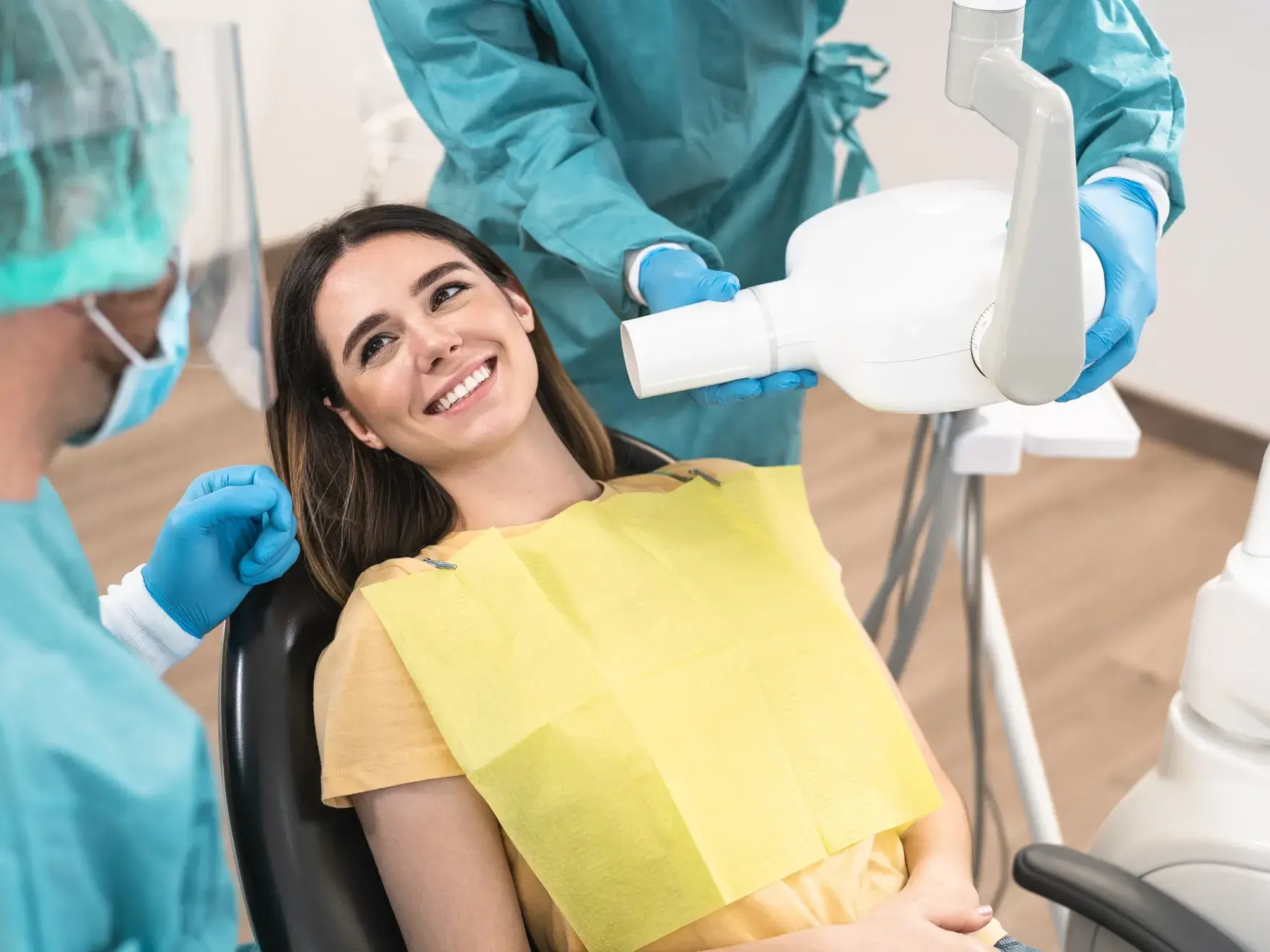 Smiling woman in dental chair with dentist and assistant preparing X-ray machine.