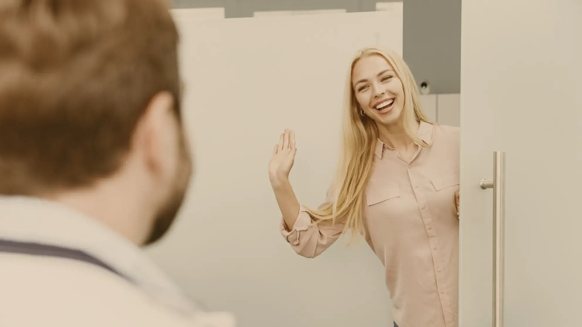 Smiling blonde woman waving and leaning out from behind an open door towards a man.