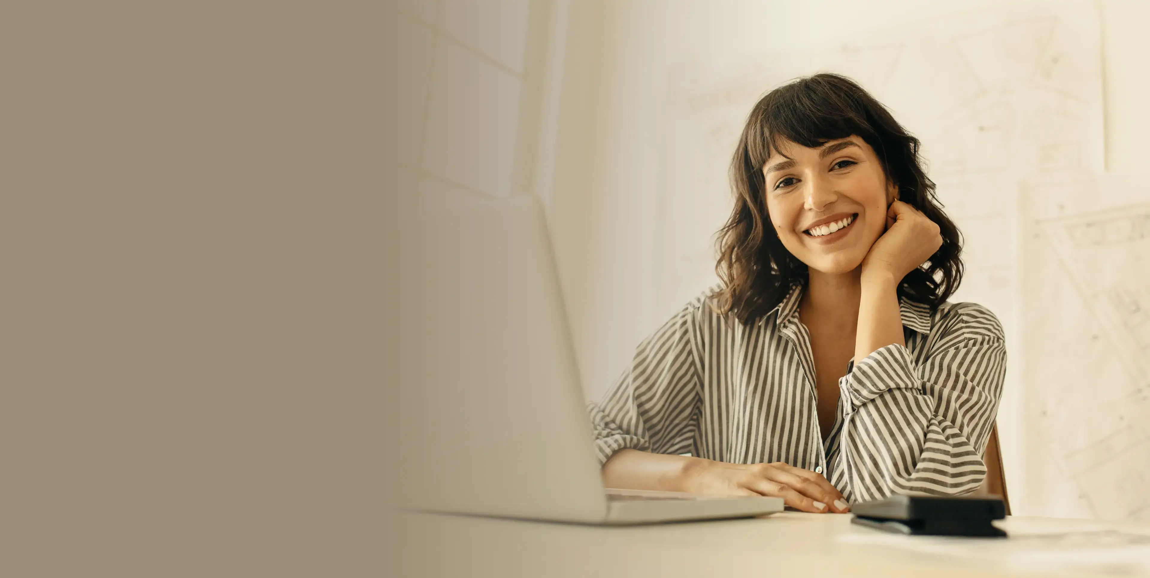 Smiling woman with dark hair in a striped shirt sitting at a desk with a laptop and phone.