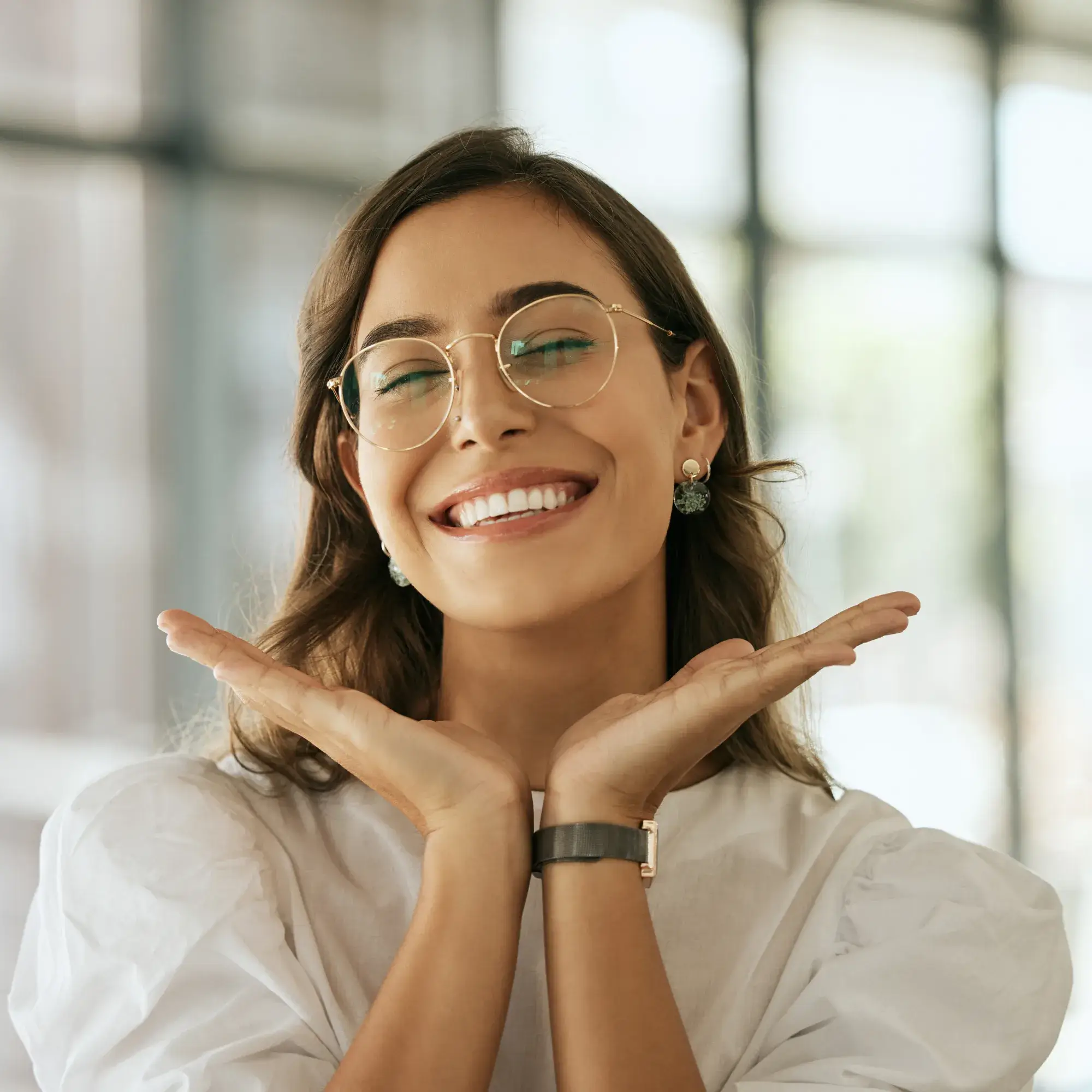 Smiling woman with glasses and earrings, hands gently framing her face, wearing a white blouse.