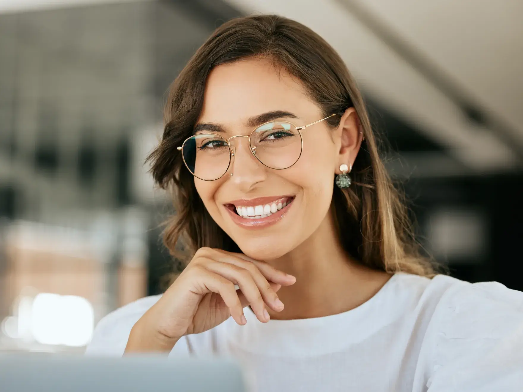 Smiling woman with glasses resting her chin on her hand in a bright indoor setting.