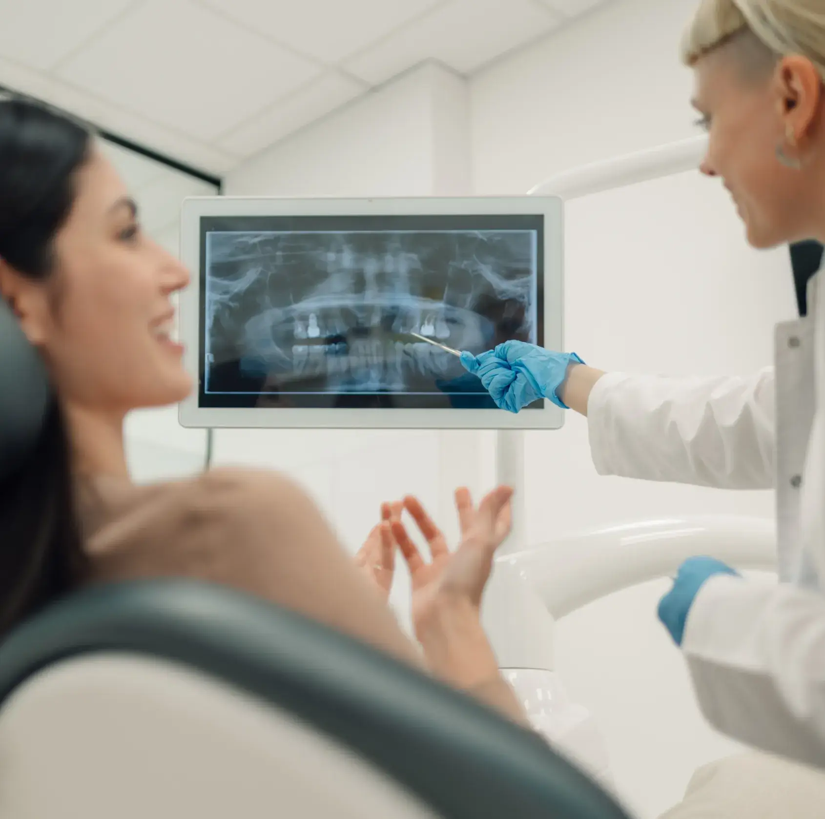 Dentist wearing blue gloves pointing at a dental X-ray on a screen while a patient smiles in a dental chair.