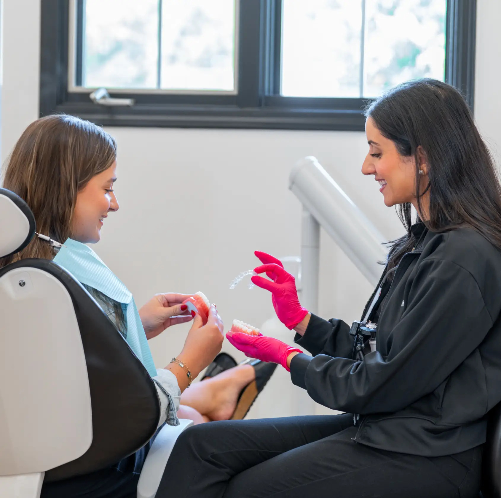 Dentist wearing blue gloves pointing at a dental X-ray on a screen while a patient smiles in a dental chair.