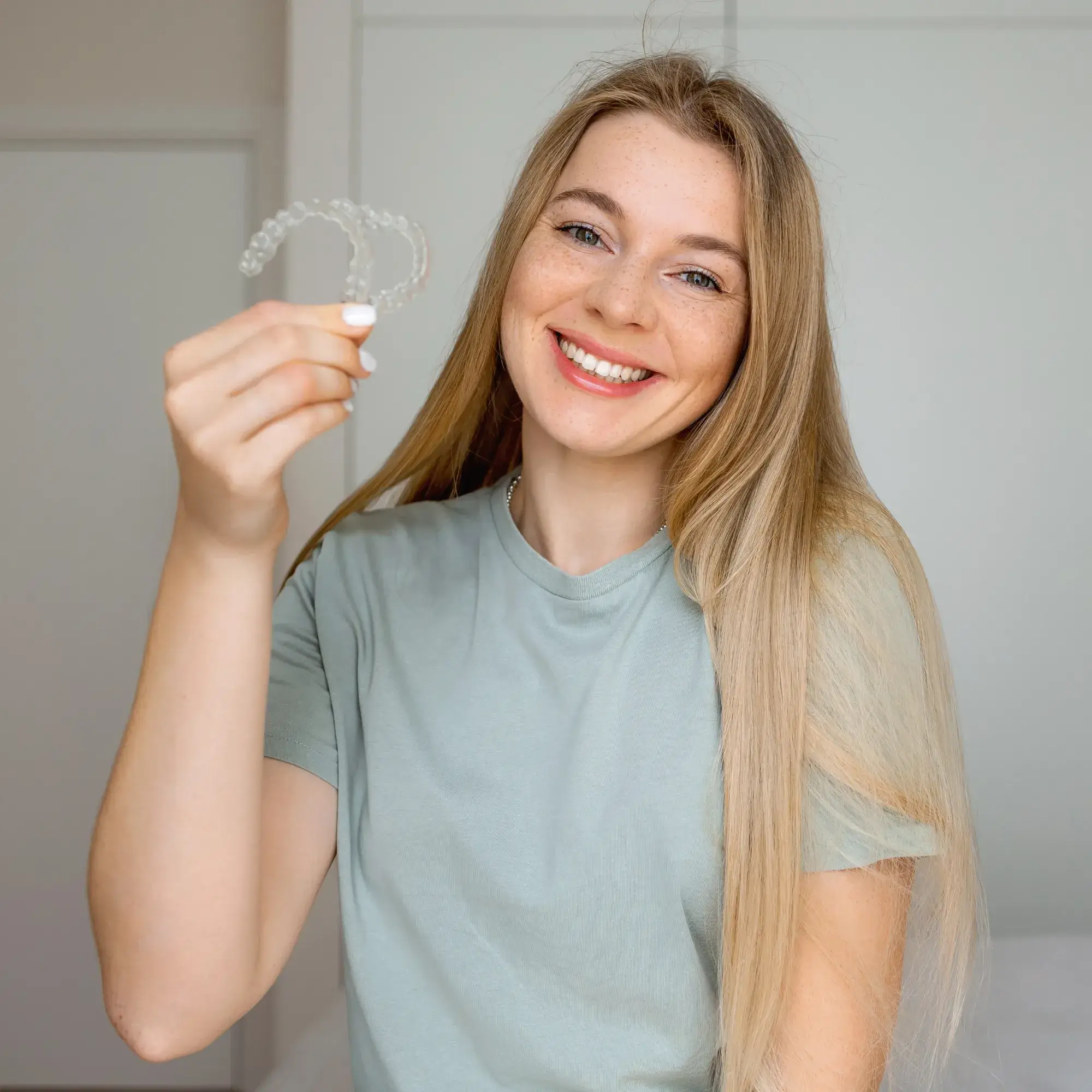 Smiling woman with long blonde hair holding clear dental aligners in her hand.