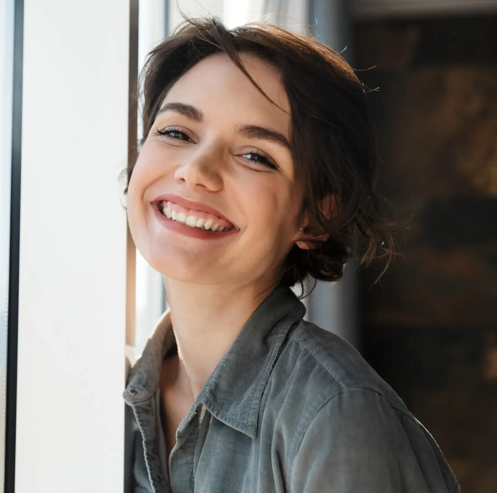 Young woman with short dark hair smiling warmly by a window.