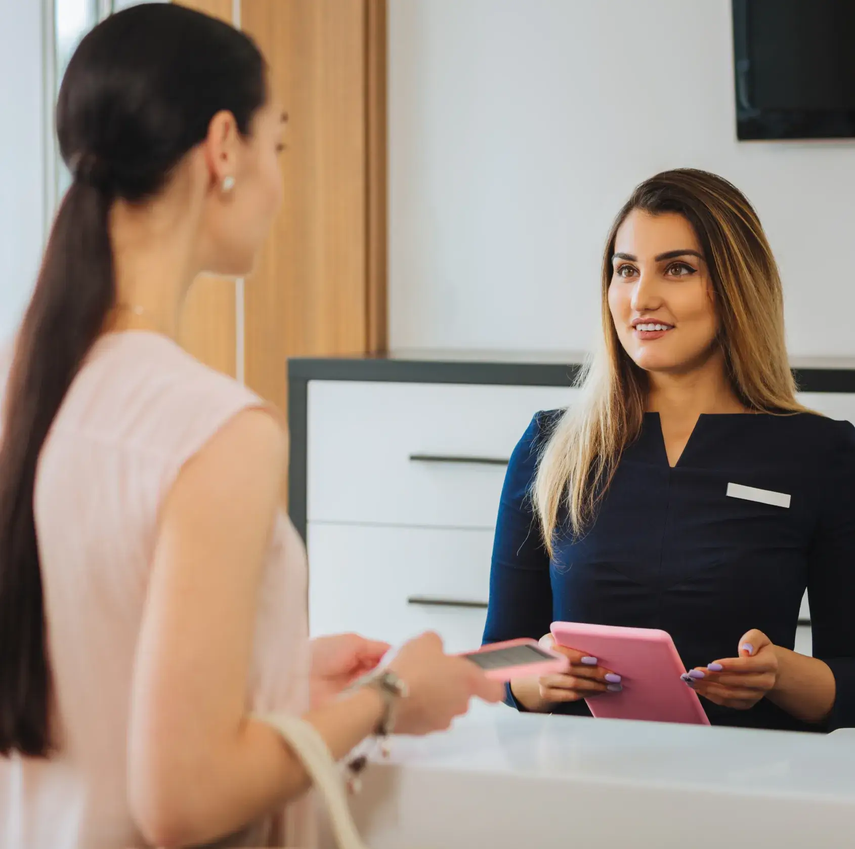 Receptionist in navy uniform holding a pink clipboard and talking to a woman with long dark hair.