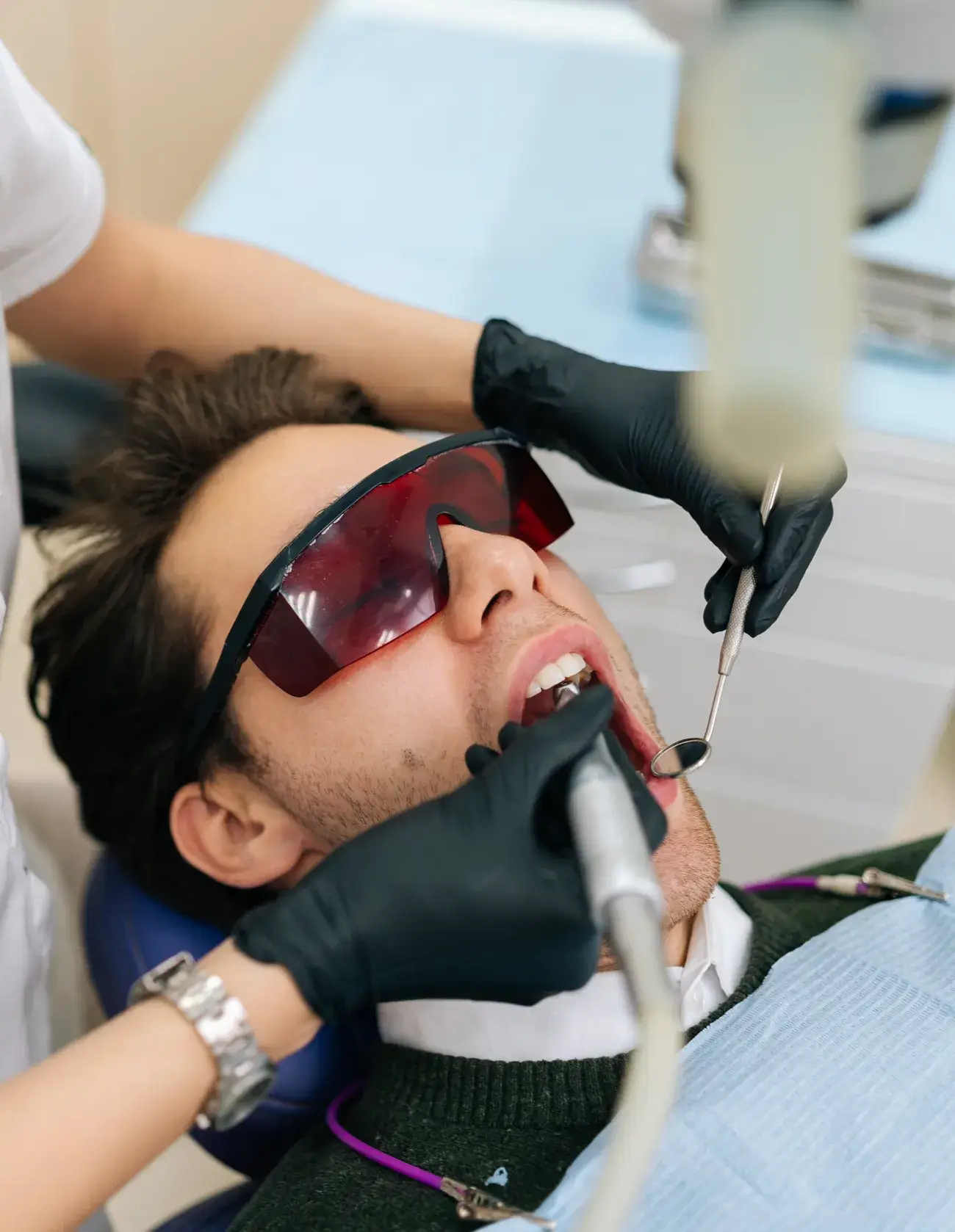 Man wearing protective red glasses receiving dental treatment with dental instruments held by a dentist in black gloves.