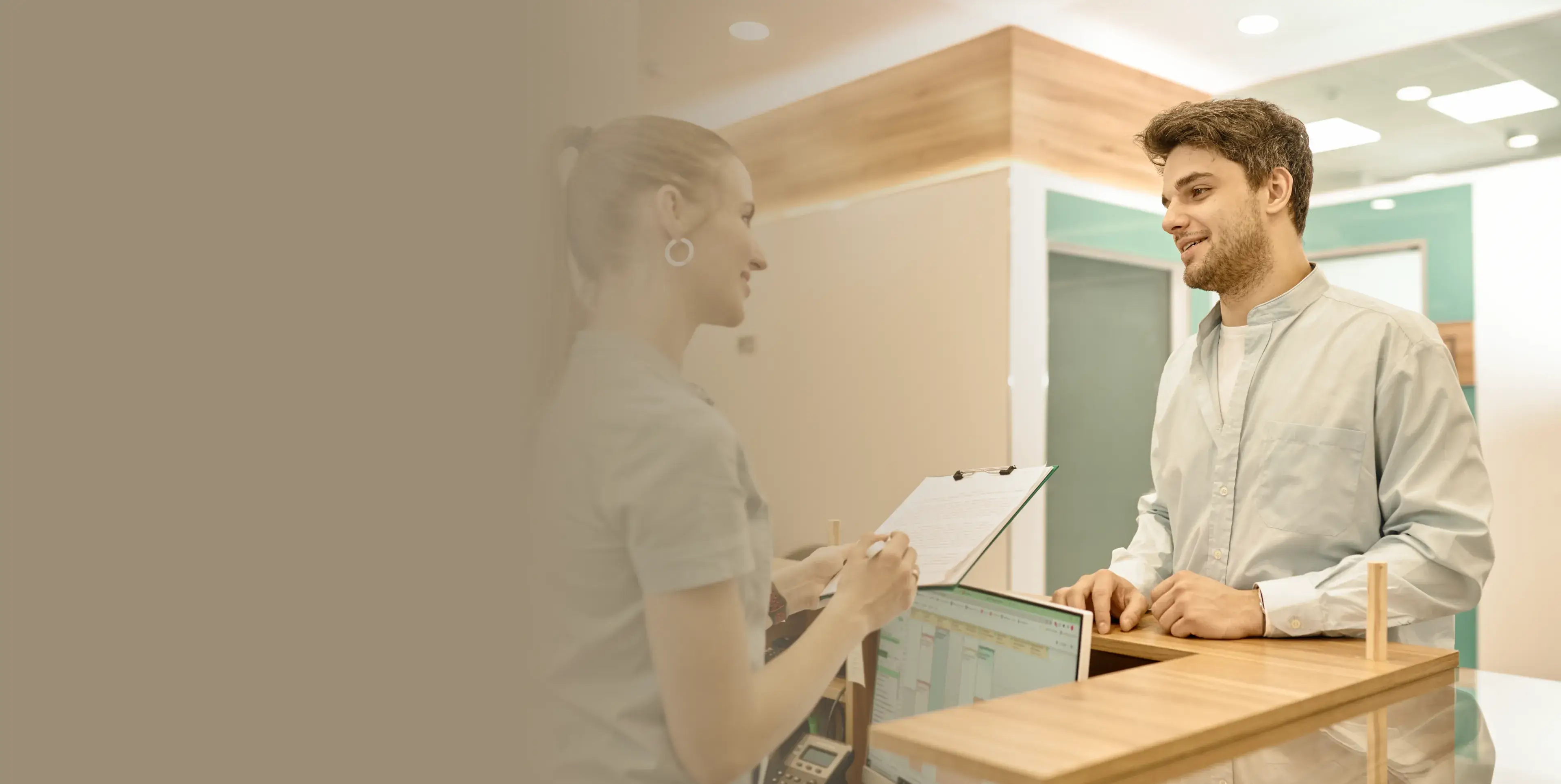 A receptionist with a clipboard speaking to a young man standing at a wooden front desk in a bright, modern office.