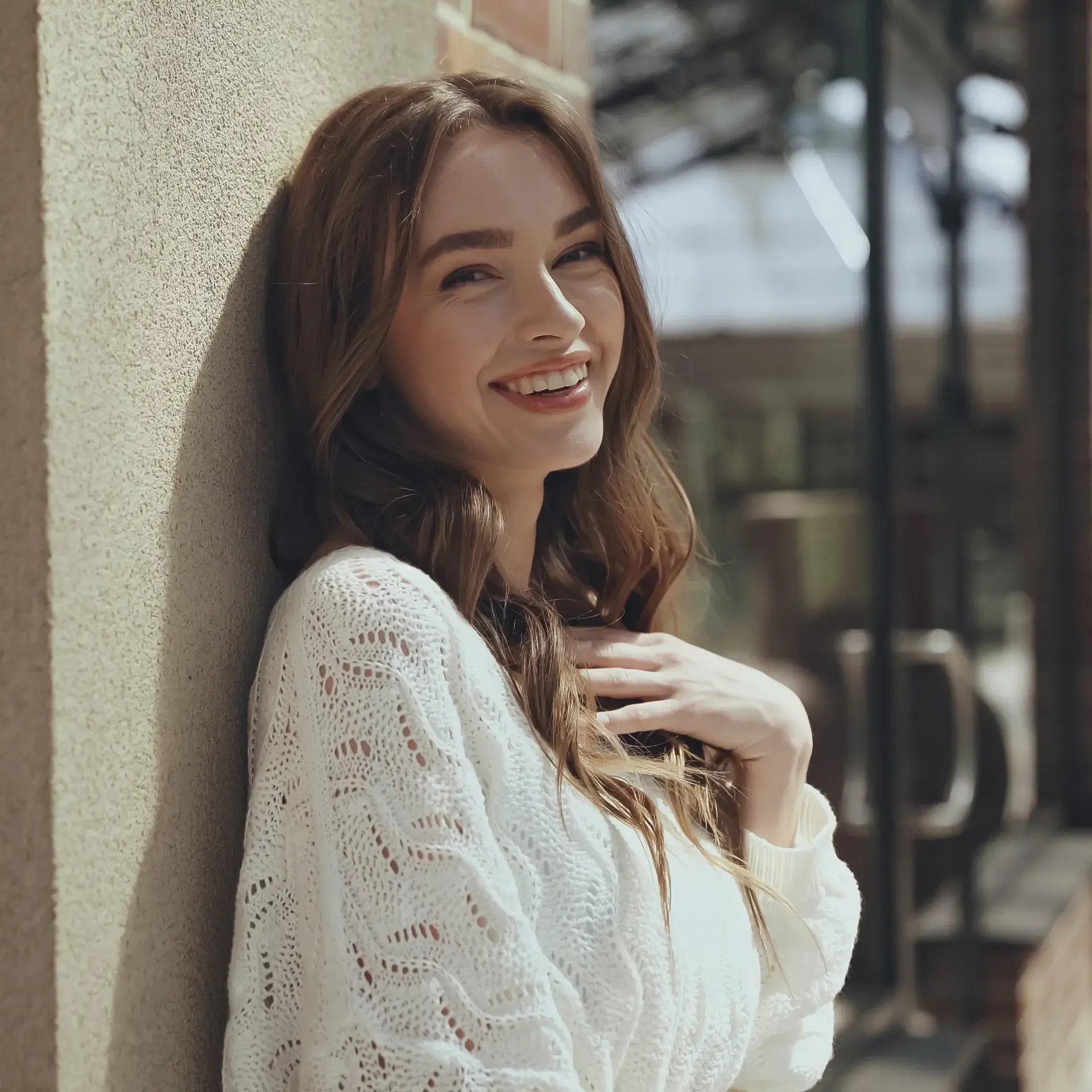 Smiling young woman in a white knitted sweater leaning against a wall with her hand on her chest.