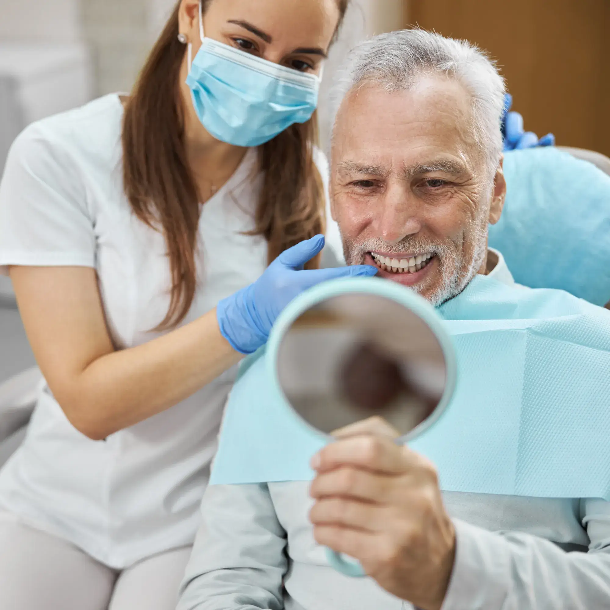 Dentist wearing mask and gloves examining elderly man's teeth as he looks in a handheld mirror.