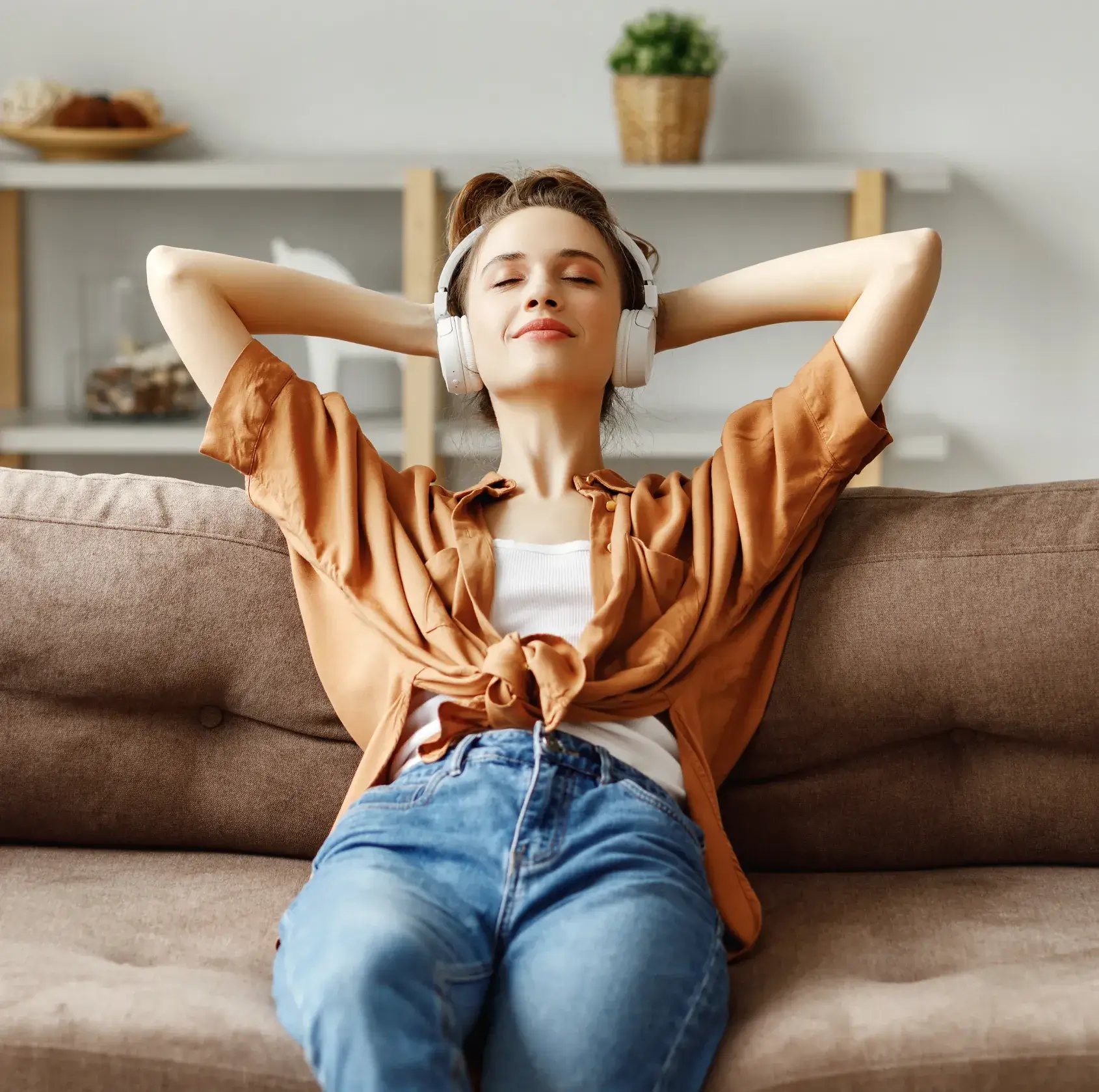 Woman relaxing on a couch with eyes closed, wearing headphones and a brown shirt over a white top.