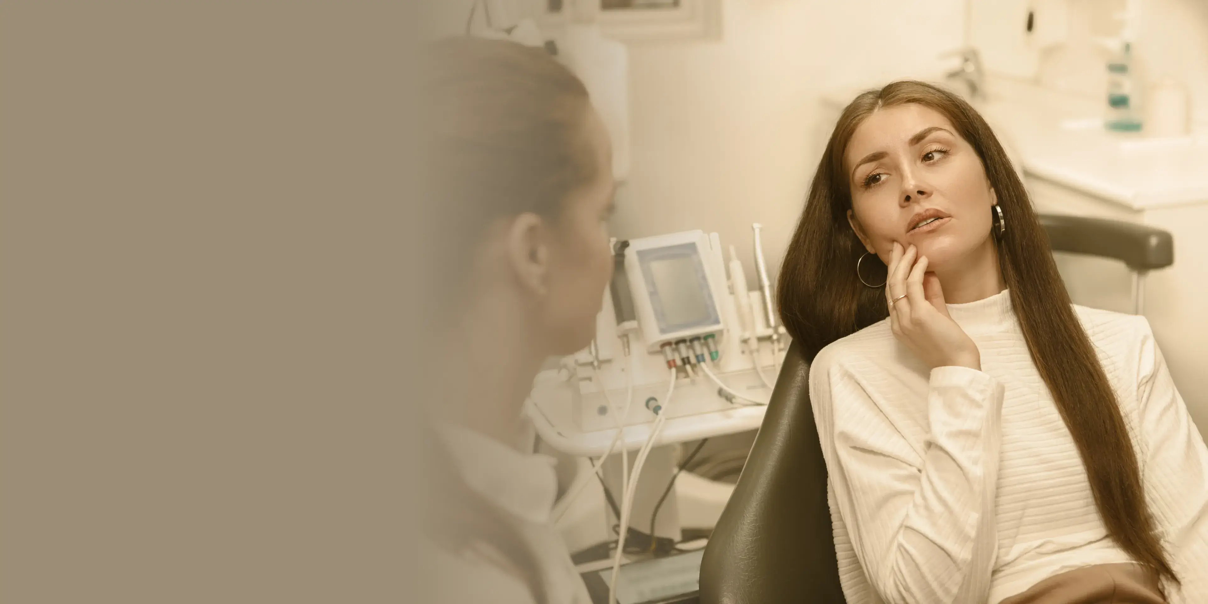 Woman sitting in a dental chair, touching her cheek, while a dental professional talks to her in a clinic.