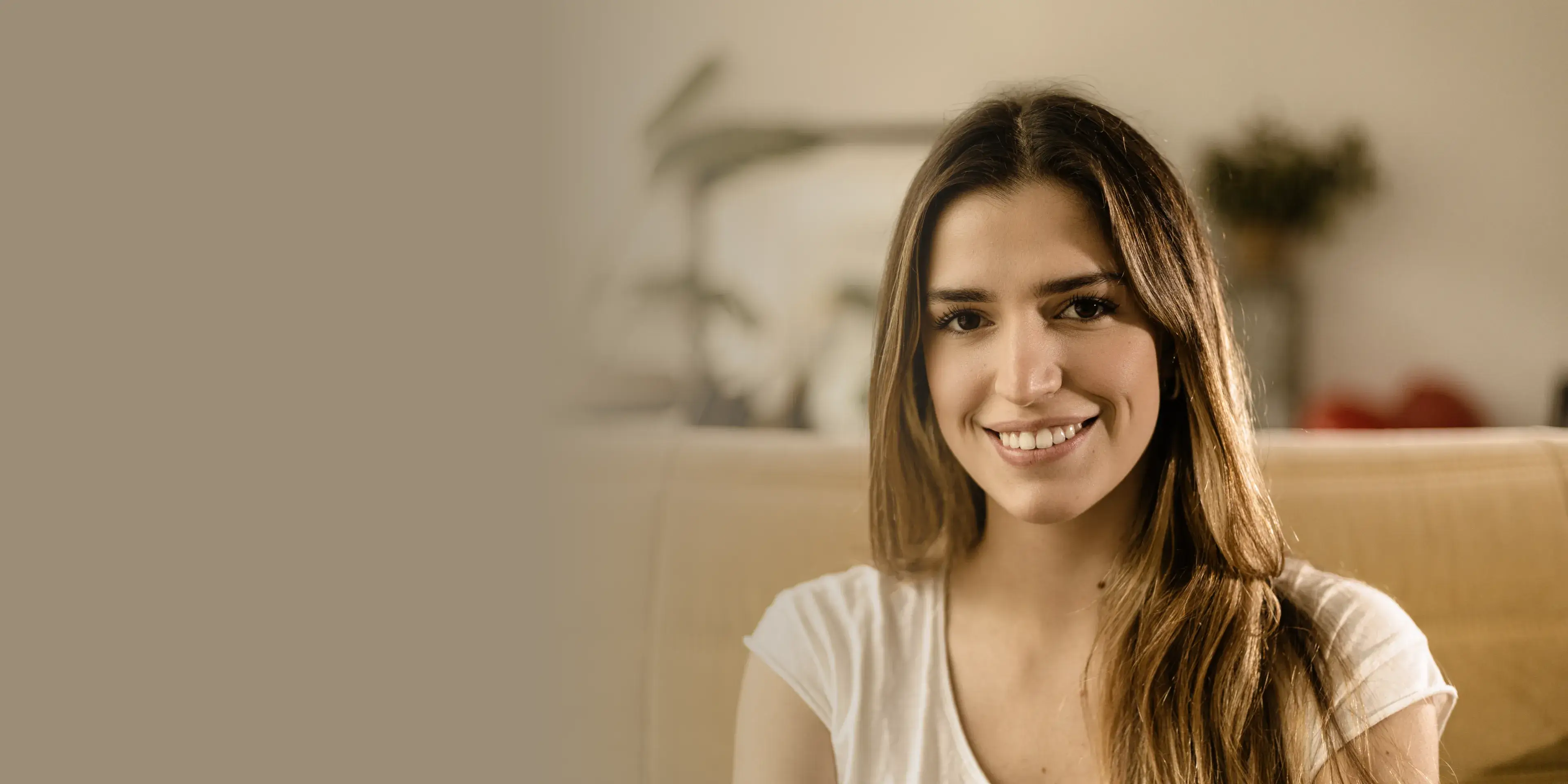 Smiling young woman with long brown hair sitting on a beige couch in a warmly lit room.