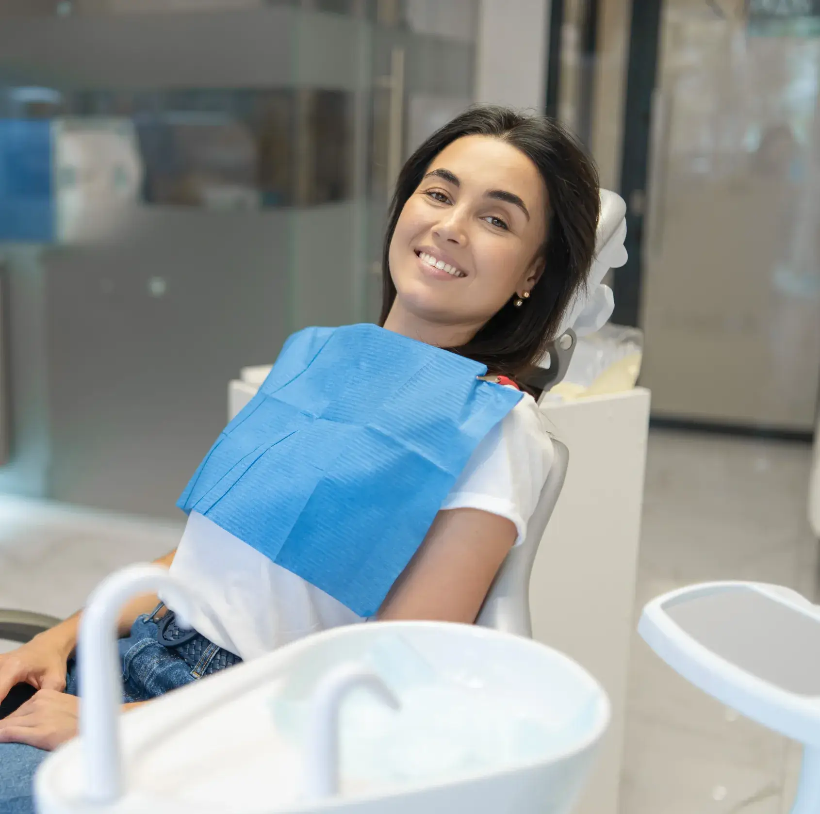 Smiling woman sitting in a dental chair wearing a blue dental bib in a modern dental clinic.