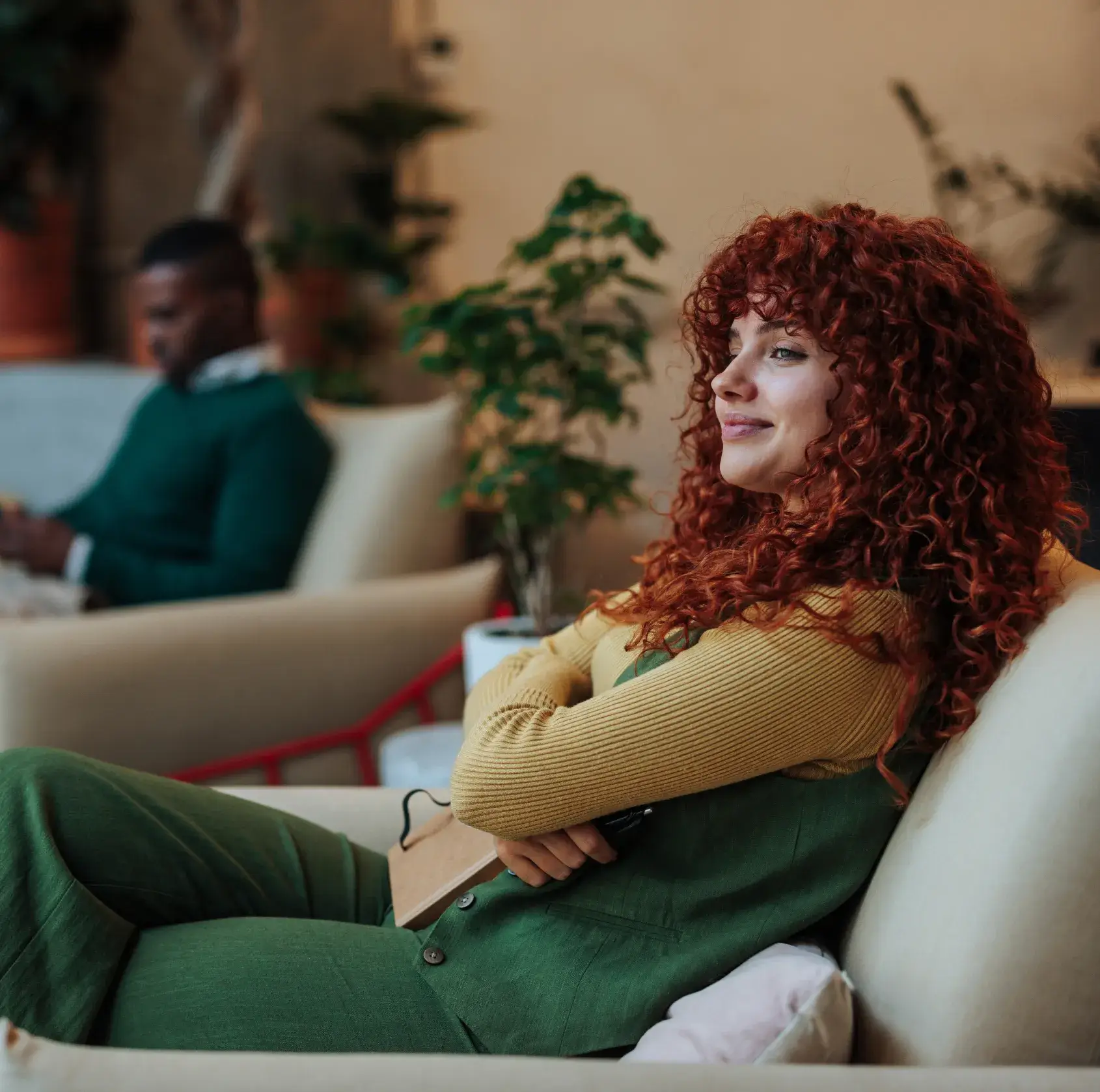Smiling woman with curly red hair sitting on a couch with arms crossed, man blurred in the background.